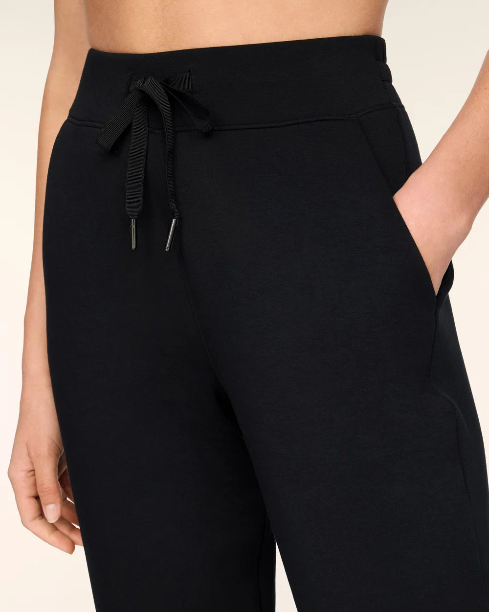 Close-up of a person wearing black drawstring sweatpants with side pockets, shown against a light beige studio background.