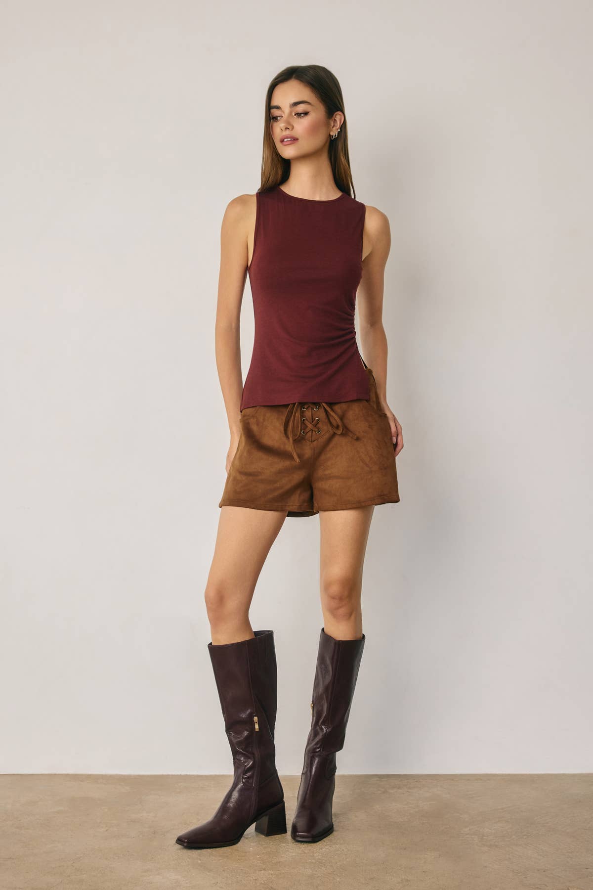 Woman wearing a burgundy sleeveless top and brown shorts with knee-high boots against a plain background