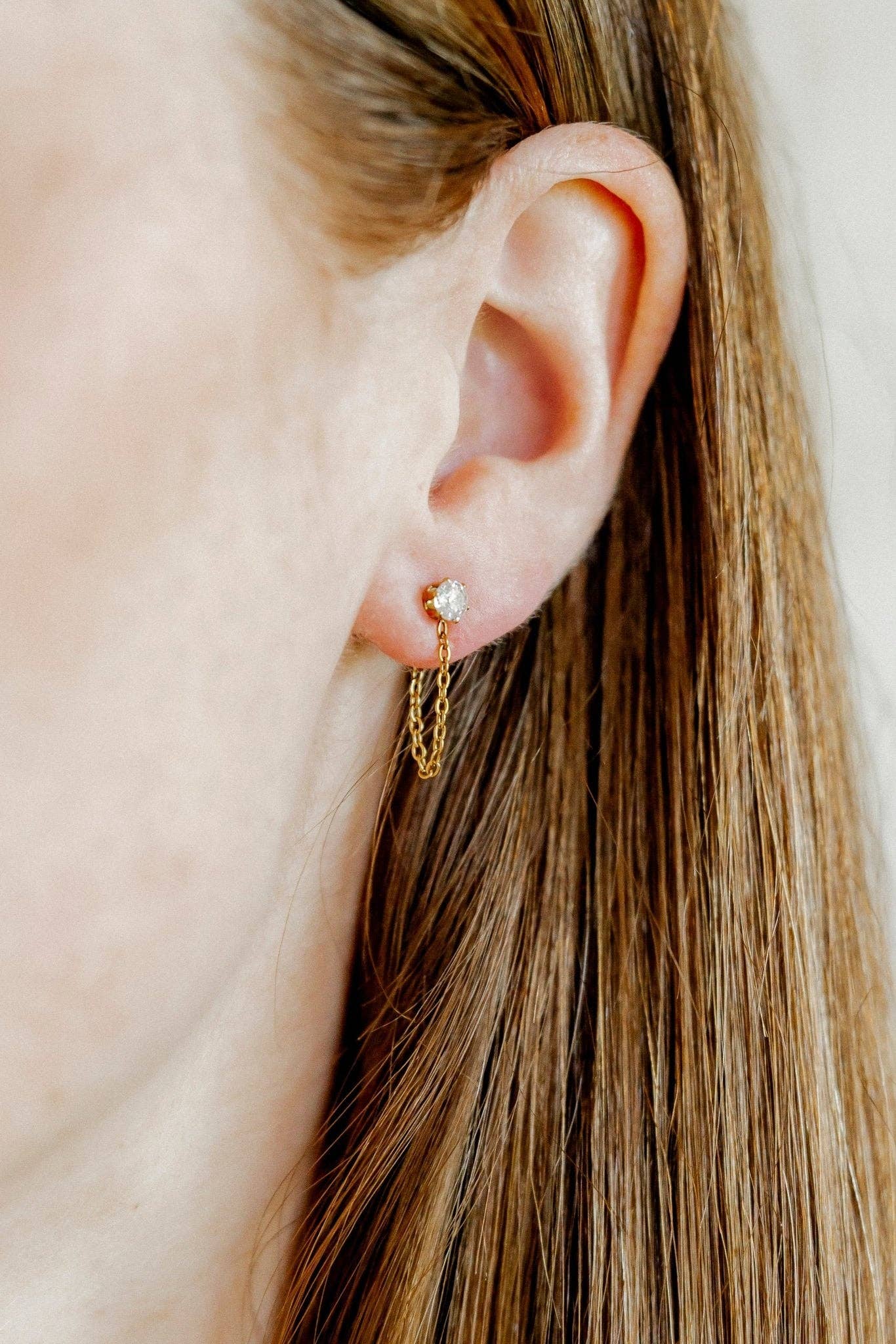 A close-up of a person's ear wearing a delicate gold dangle earring with a small clear stone and a short chain drop, against a soft light background.