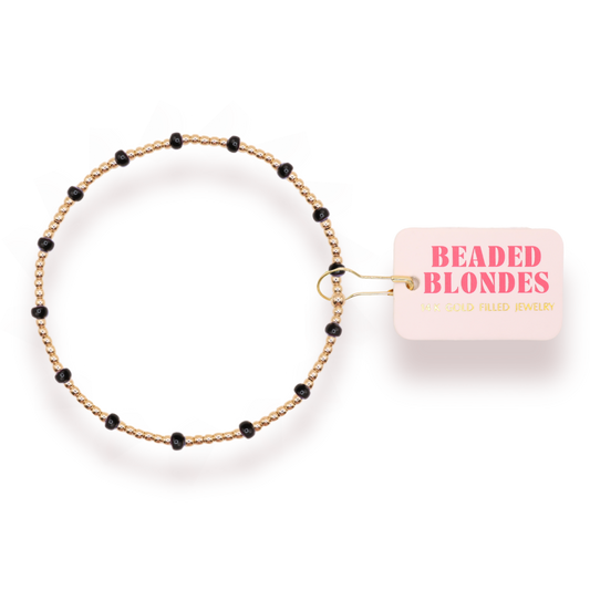 A delicate beaded bracelet with a gold-toned twisted wire base and small black beads spaced along the band, shown next to a light pink paper tag that reads 'BEADED BLONDES'.