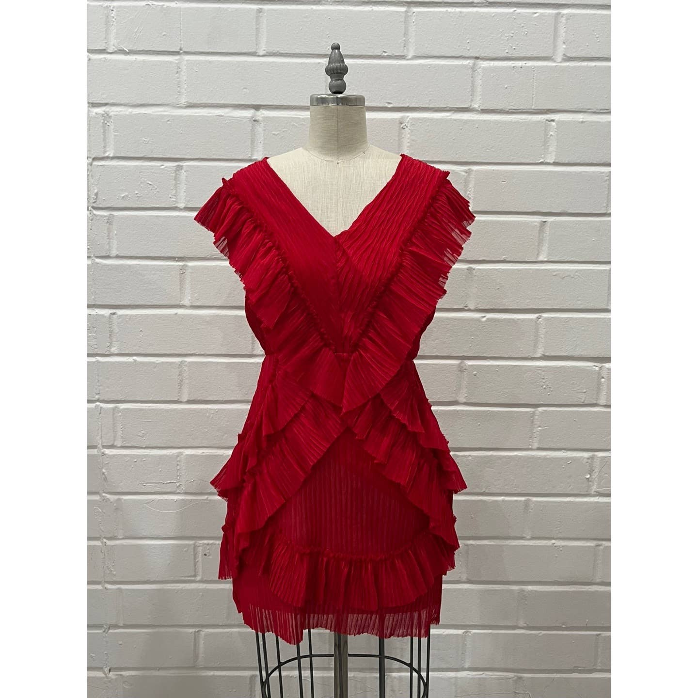 A bright red pleated ruffle dress displayed on a dress form mannequin against a white painted brick wall background.