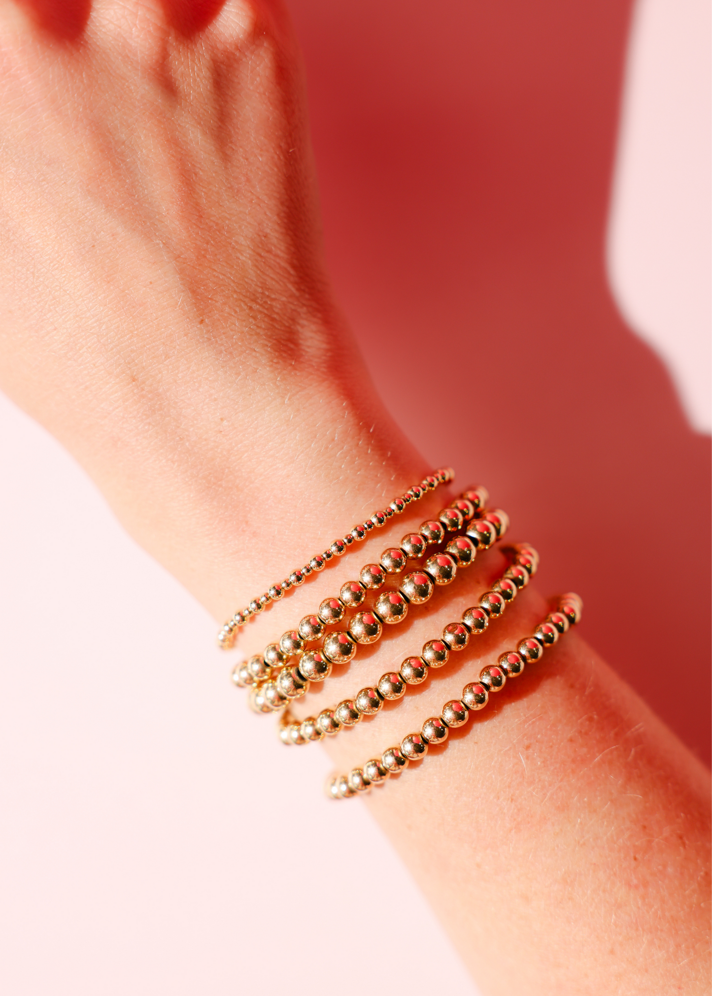 Close-up of a wrist wearing multiple gold beaded bracelets on a pink background