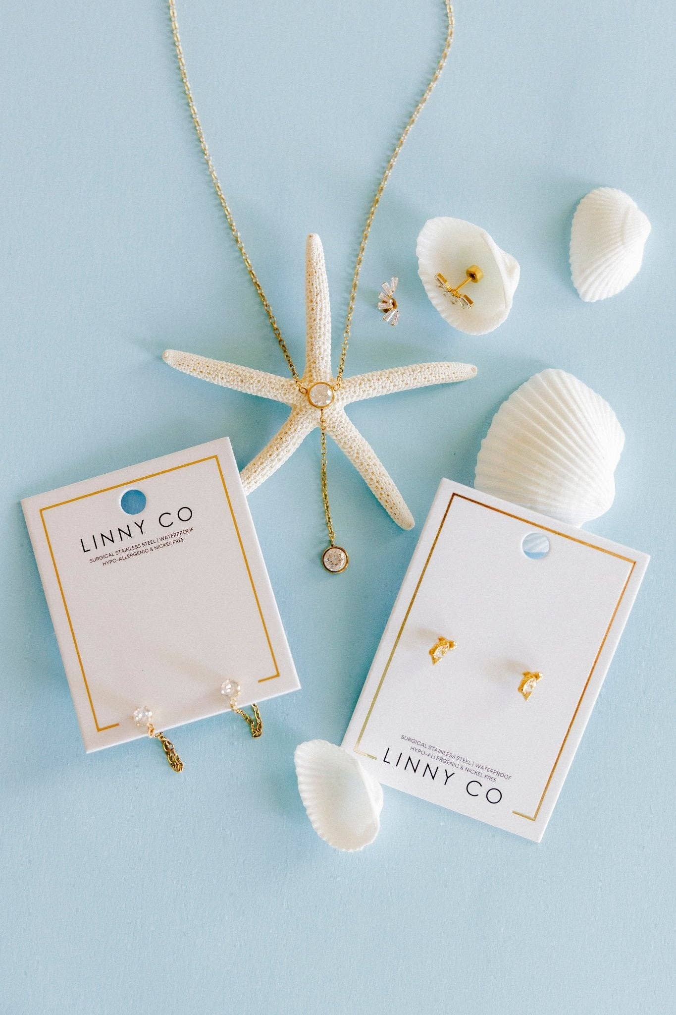 A flat-lay of jewelry from the brand LINNY CO on a light blue background, featuring a gold necklace with a starfish pendant and matching gold stud earrings on white display cards, styled with white seashells.