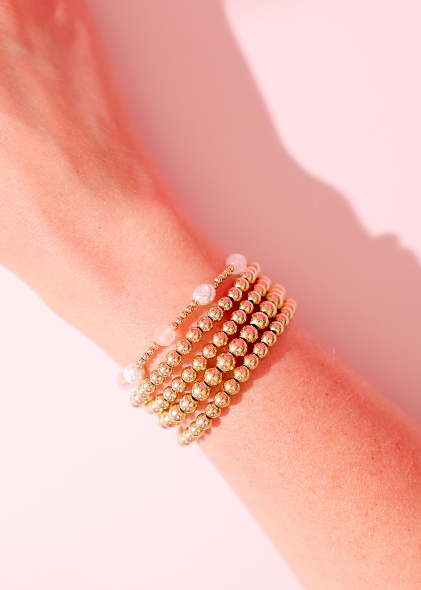 Gold beaded bracelets on a wrist against a pink background