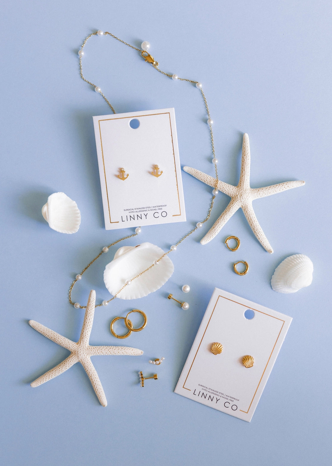 Gold jewelry set with starfish, shells, and 'LINNY CO' cards on a light blue background