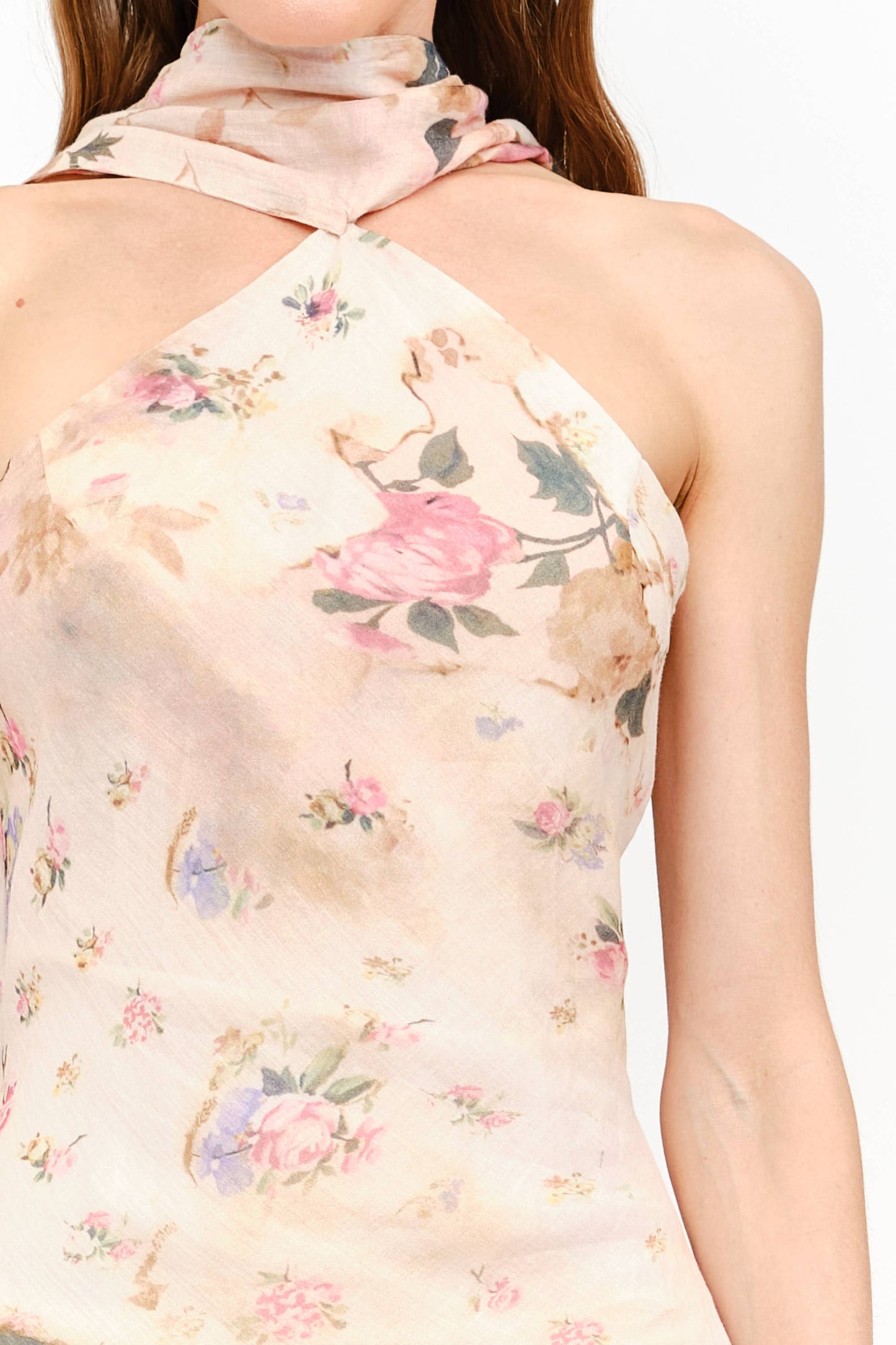 A close-up of a woman wearing a halter-neck top with a soft blush pink floral print on a light beige base. The top features a high neck with the fabric tied behind it, and the print includes pink roses, green leaves, and small blue accents.