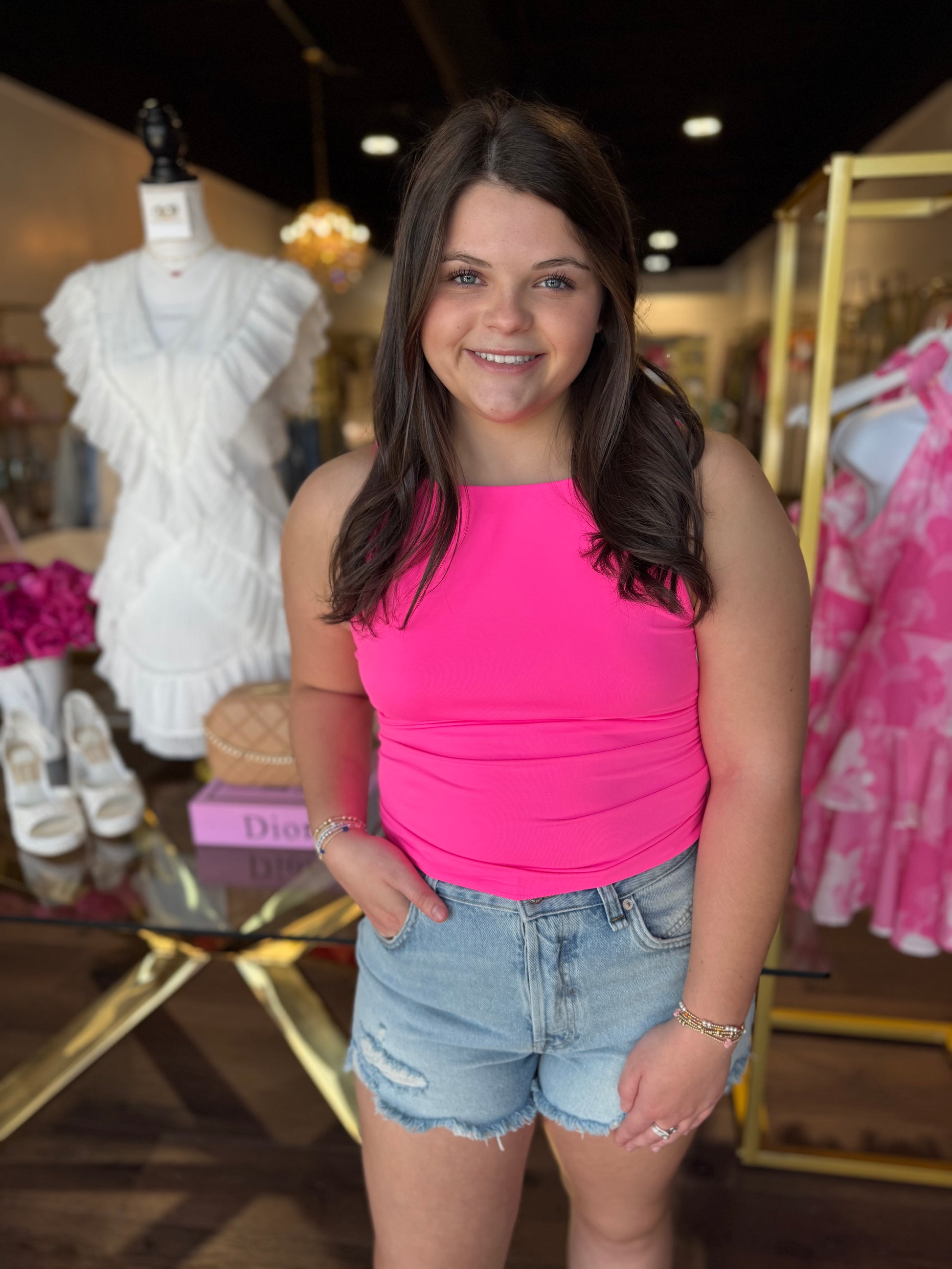 A young woman with long dark brown hair stands in a boutique wearing a bright hot pink sleeveless top and light-wash distressed denim shorts. A mannequin in a white ruffled dress and a pink patterned garment are visible in the background.