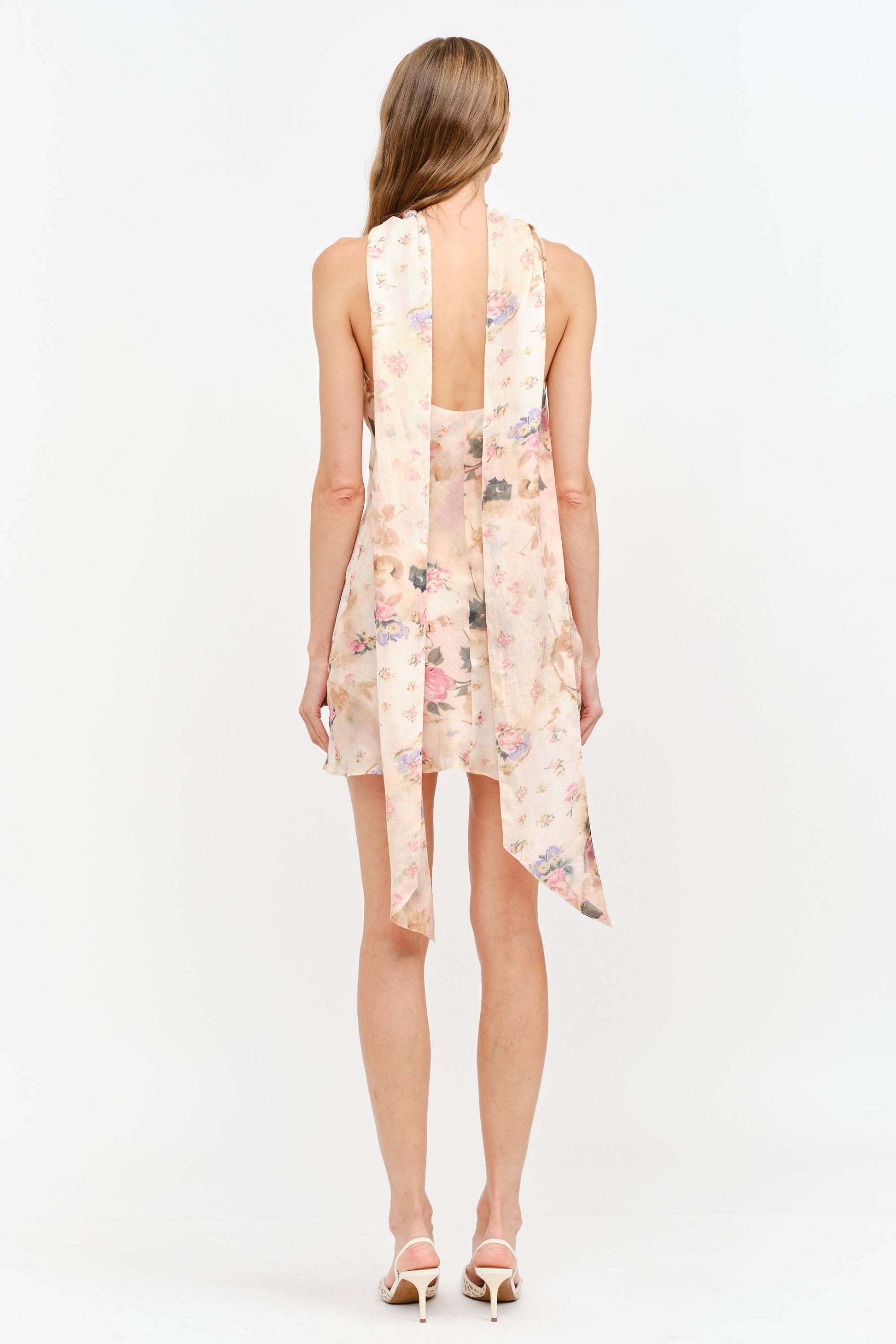 A woman shown from the back wearing a sleeveless, open-back mini dress with a long draped scarf or shawl in a light peach-pink floral print, paired with beige high-heeled sandals, against a plain white studio background.