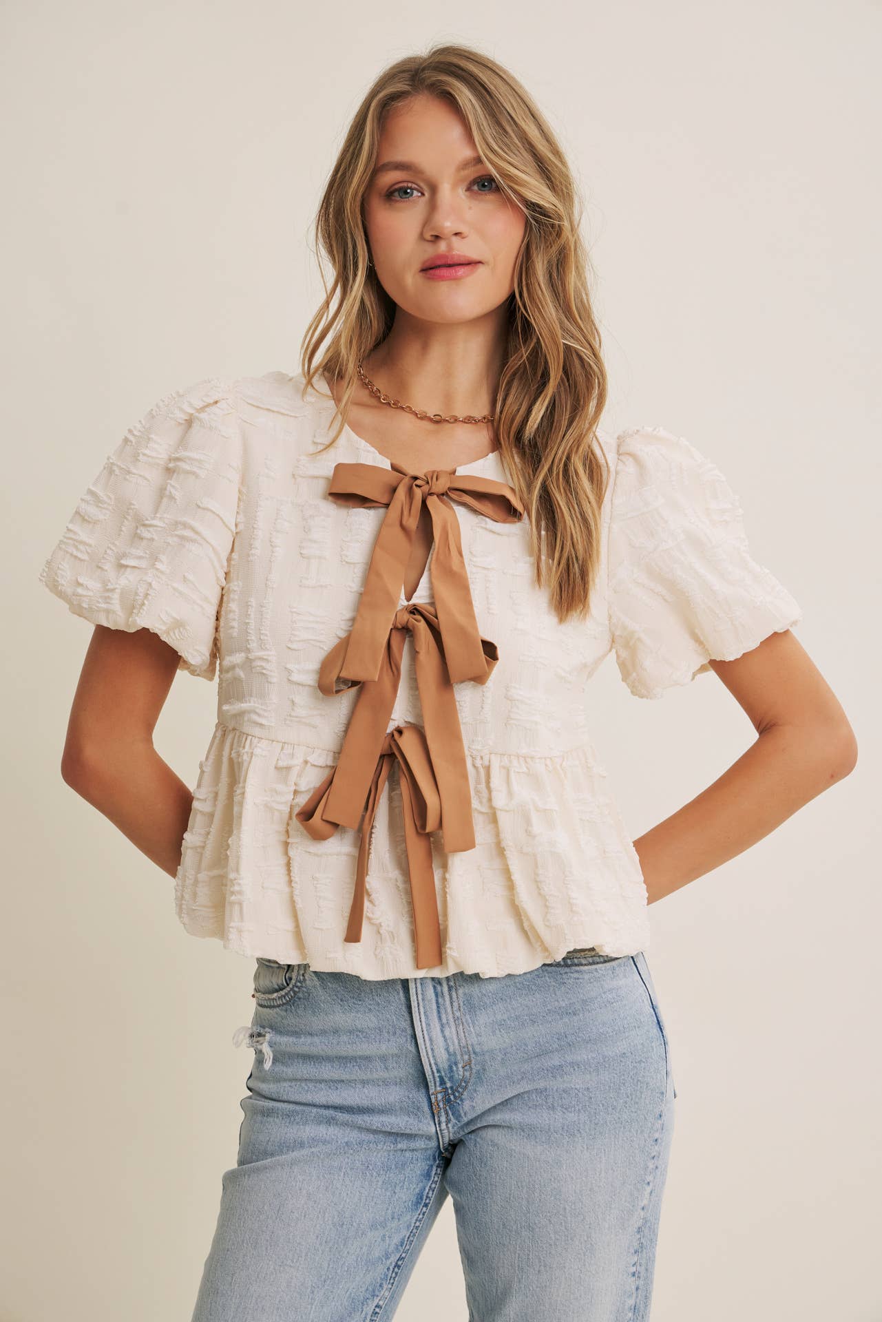 Woman wearing a white blouse with ruffled sleeves and a brown bow tie, paired with blue jeans on a plain background.
