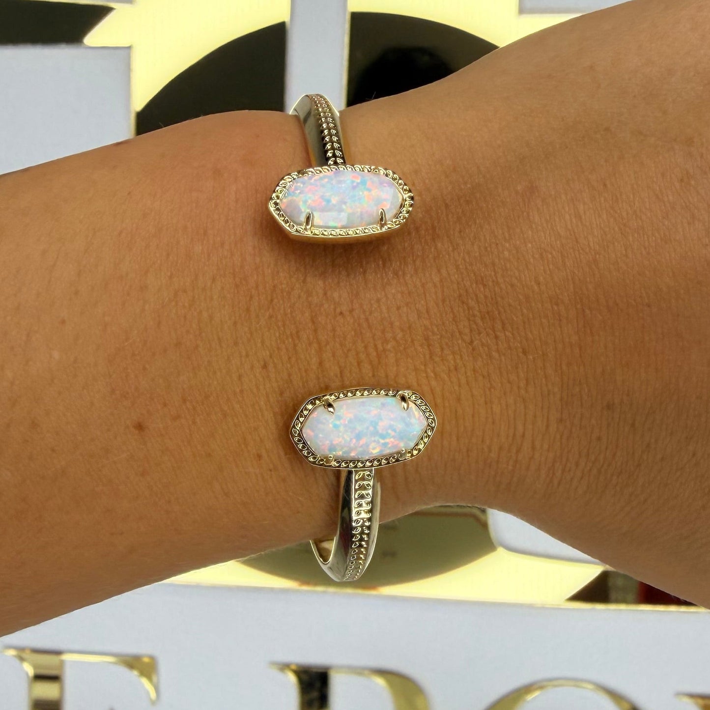 Gold bracelet with a white stone on a wrist, blurred background with decorative elements.
