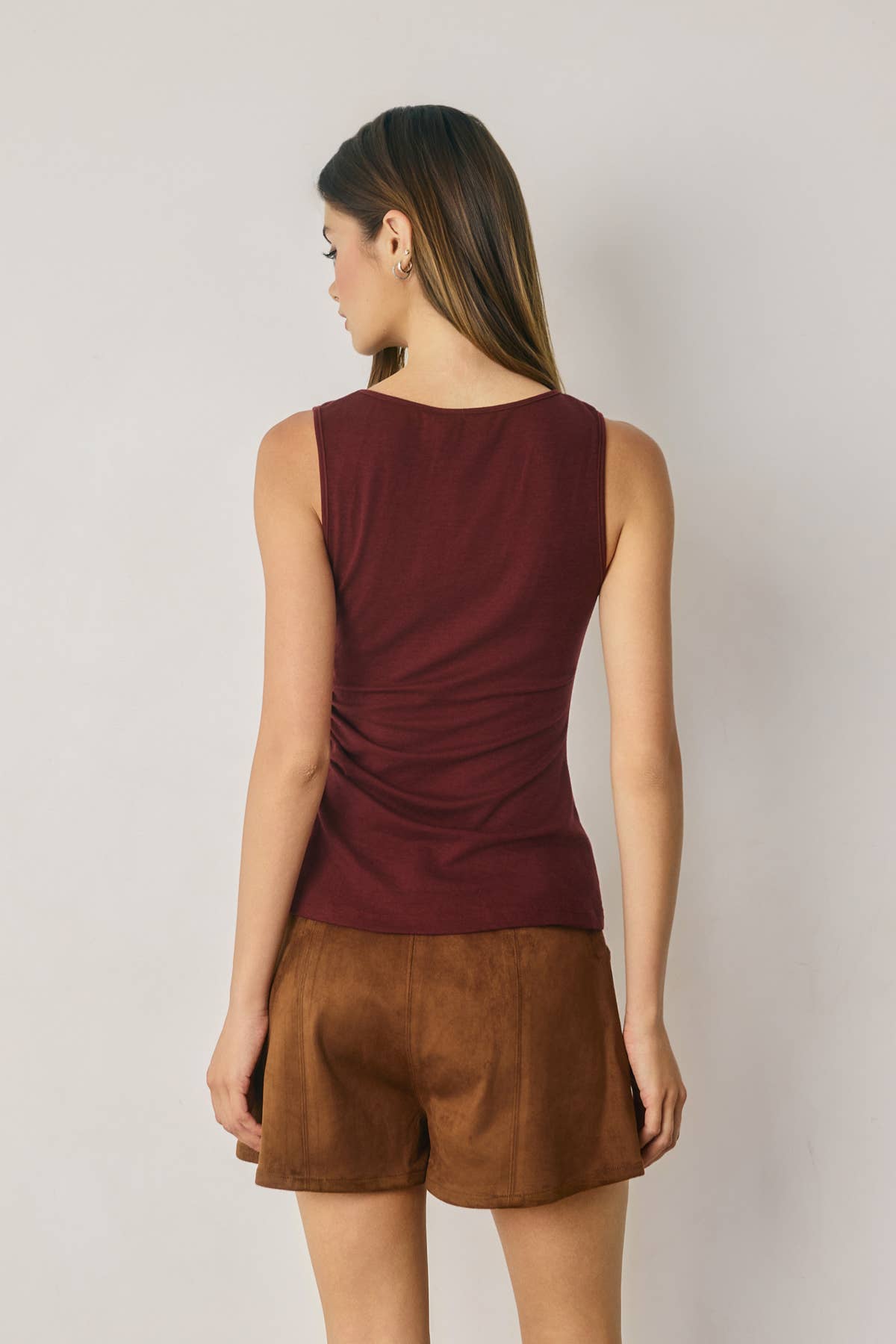 A woman shown from the back wearing a sleeveless ruched top in burgundy and brown shorts, standing against a plain light gray studio background.