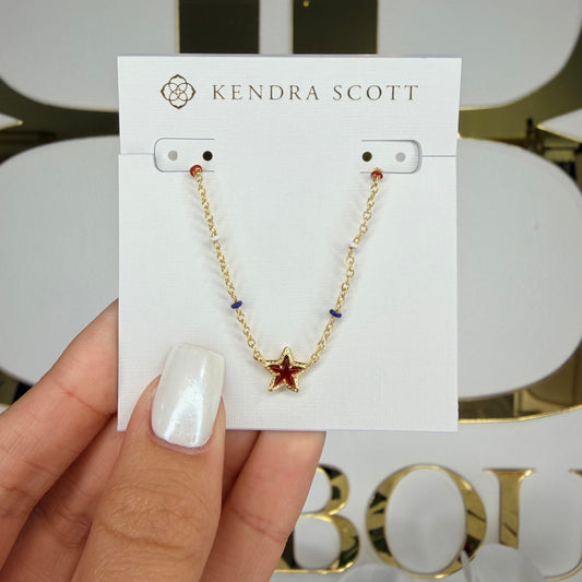 Hand holding a gold necklace with star detail on a Kendra Scott card against a blurred background.