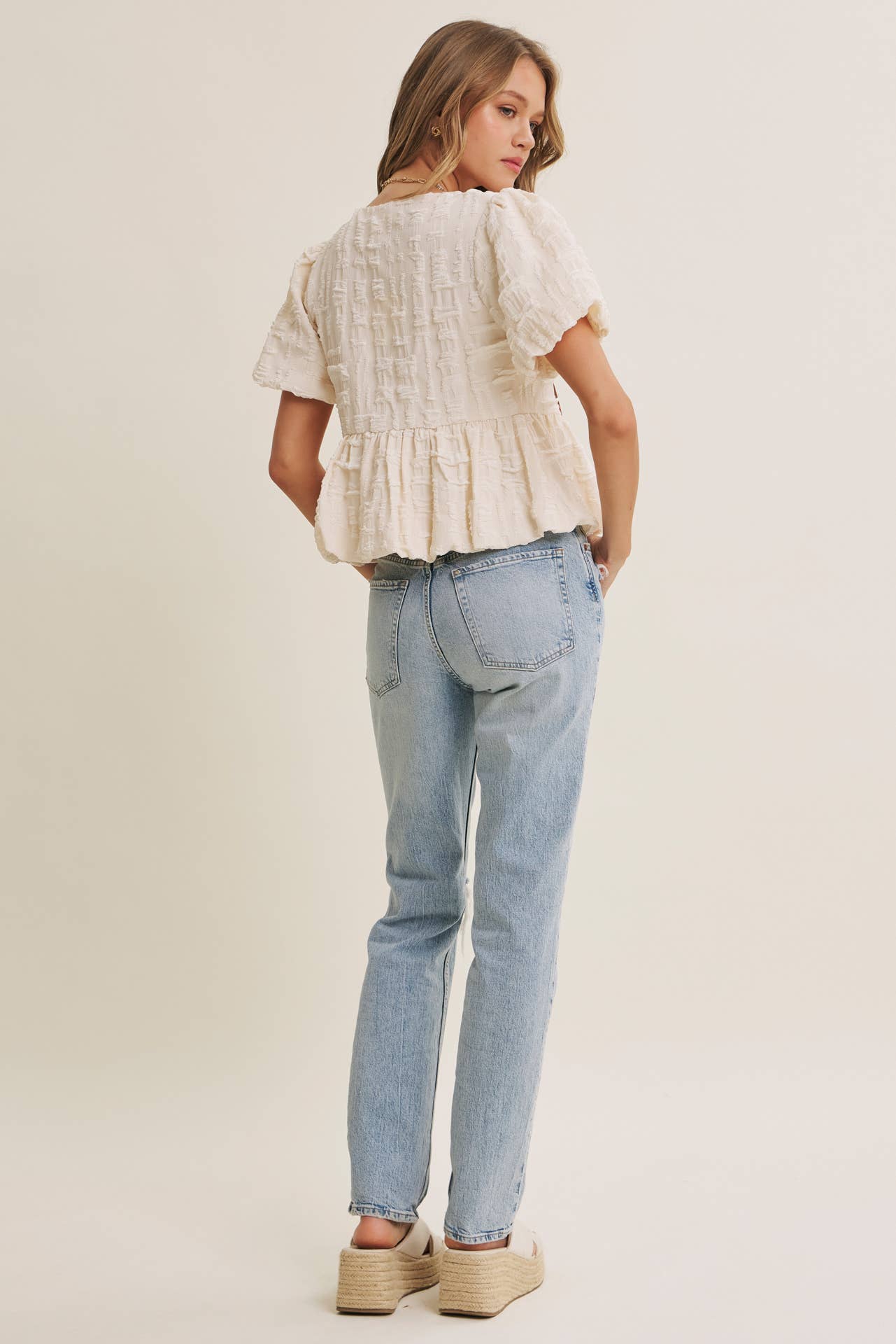 Woman wearing a white textured top and light blue jeans on a plain background
