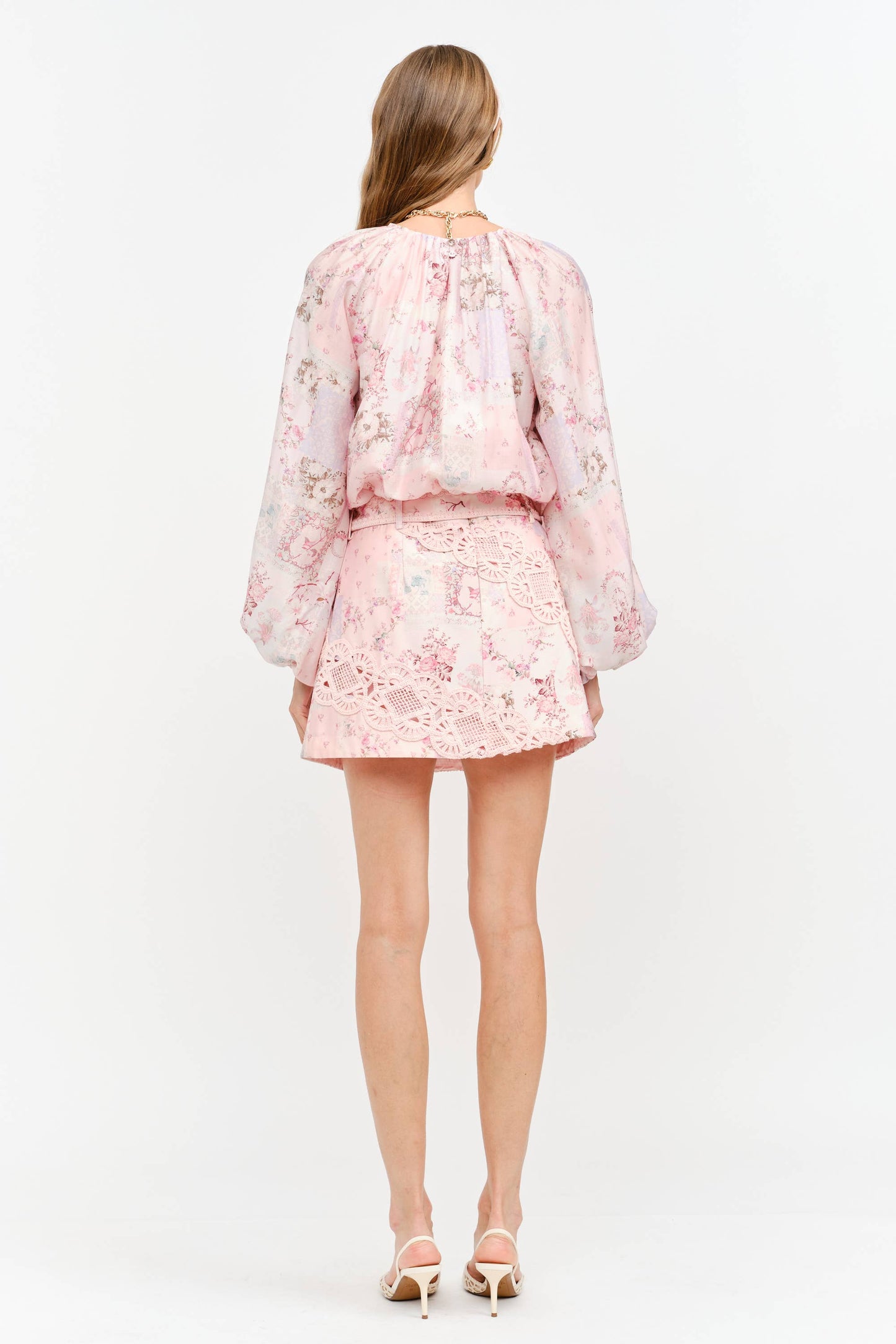 A woman viewed from the back wearing a light pink floral eyelet two-piece set with a long-sleeve blouse and matching mini skirt, paired with beige high-heeled sandals against a white studio background.