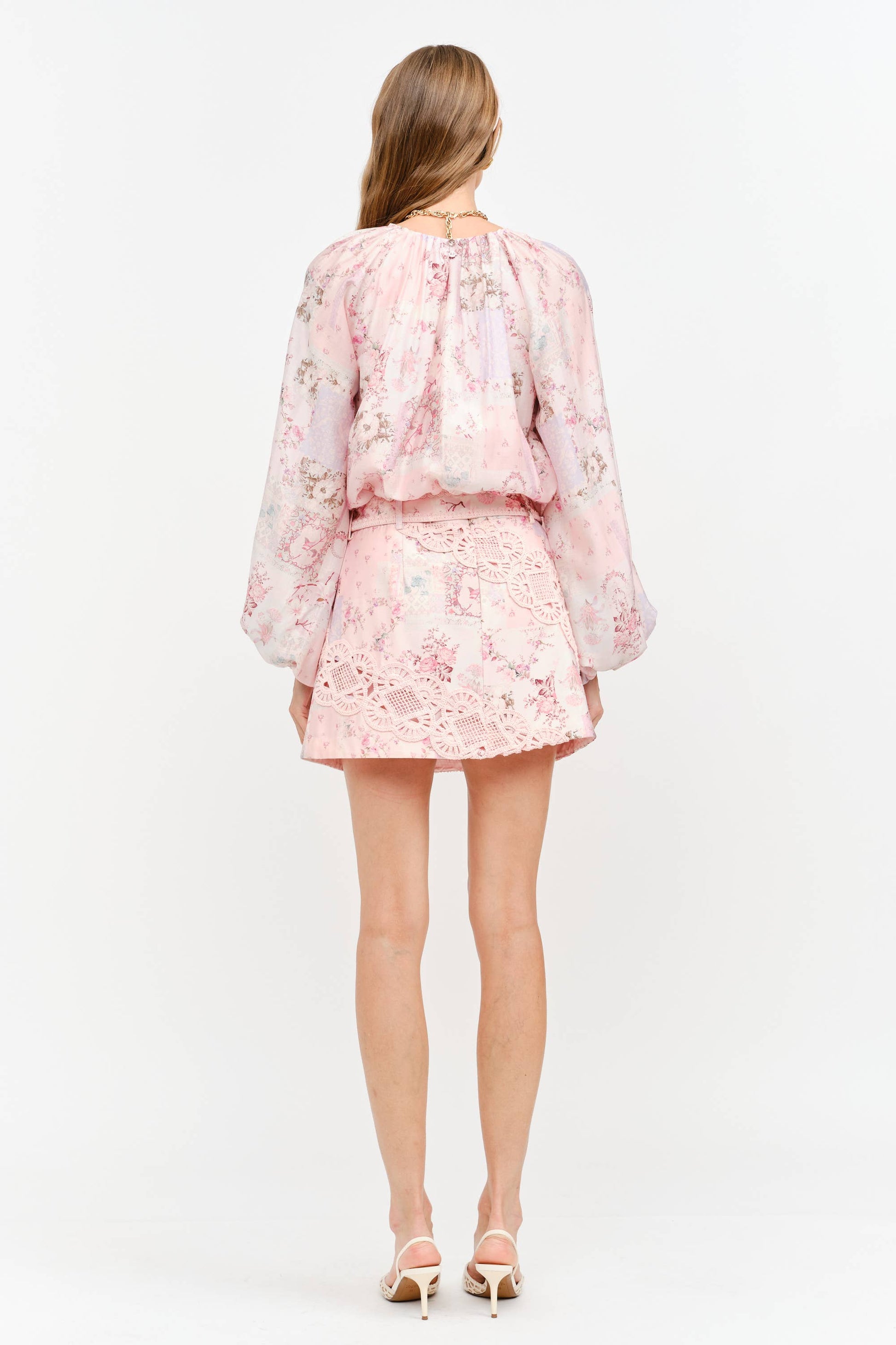A woman viewed from the back wearing a light pink floral eyelet two-piece set with a long-sleeve blouse and matching mini skirt, paired with beige high-heeled sandals against a white studio background.