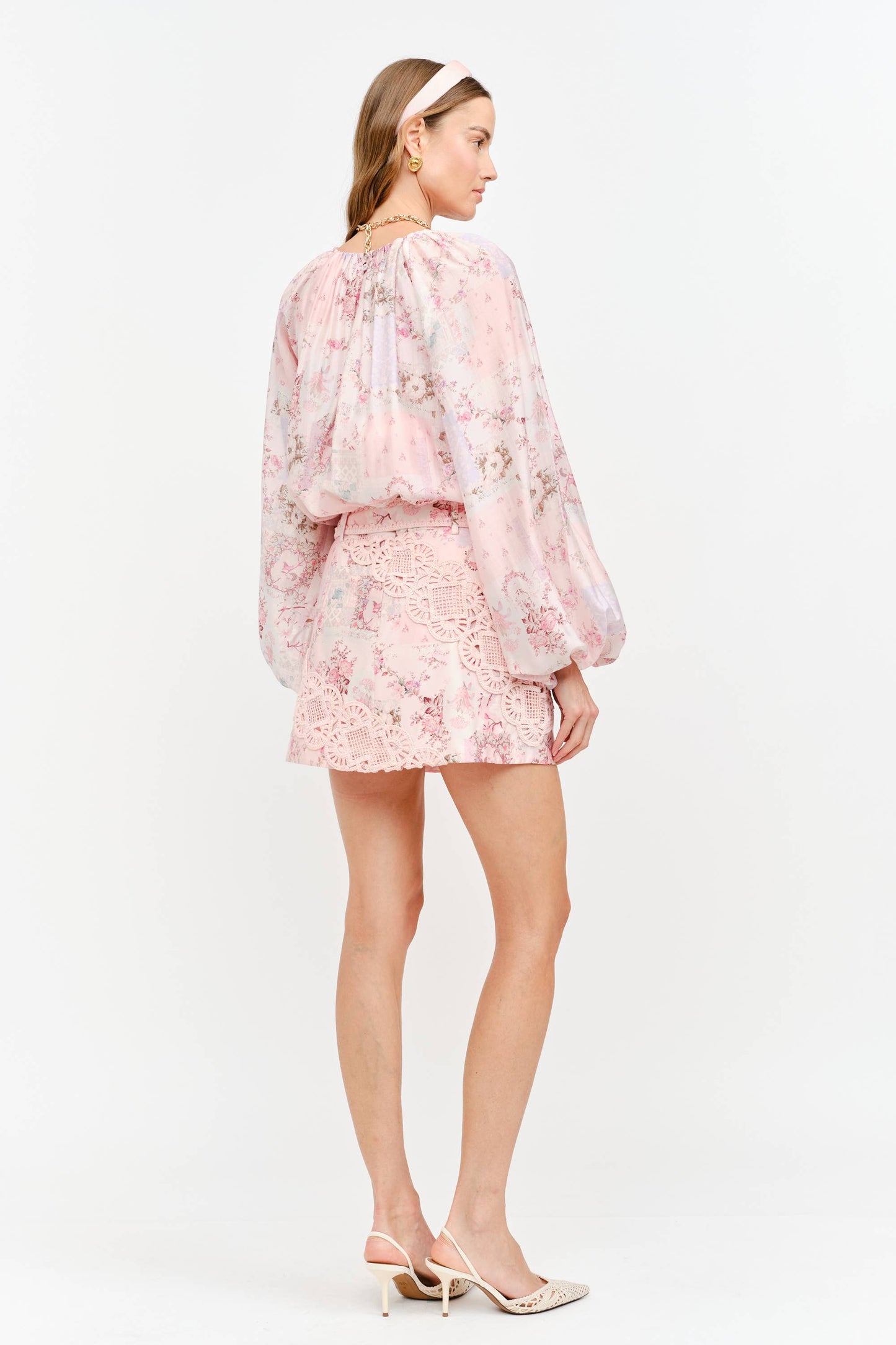 A woman modeling a light pink floral long-sleeve mini dress with a lace-trimmed neckline and short skirt, shown from the back against a plain white studio background.