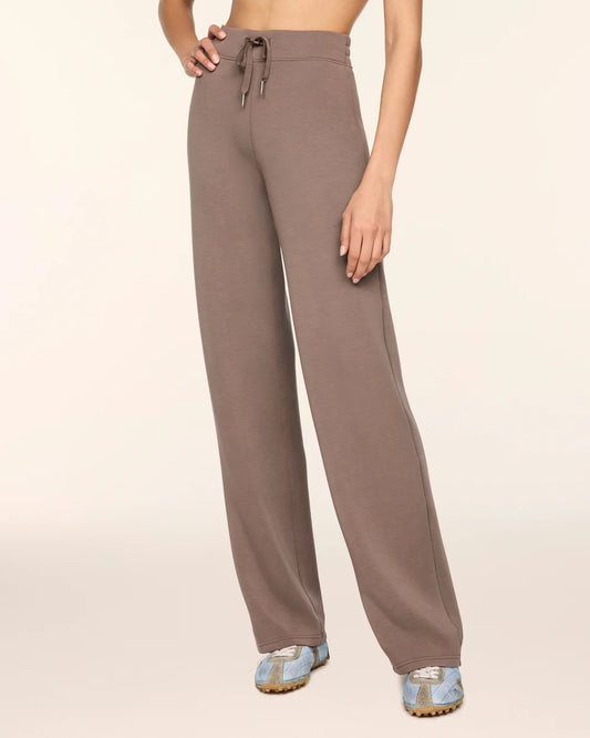 A model wearing a pair of wide-leg, high-waisted lounge pants in a muted taupe-brown color, paired with light blue sneakers, against a solid cream background.