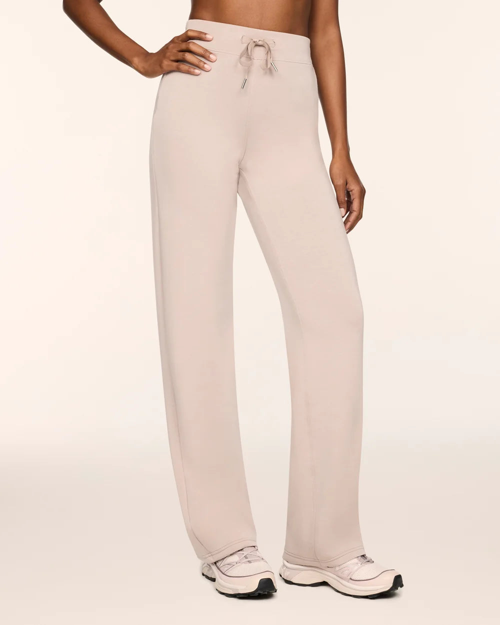 A model wearing light beige wide-leg lounge pants with a drawstring waistband, paired with white sneakers, against a solid off-white studio background.