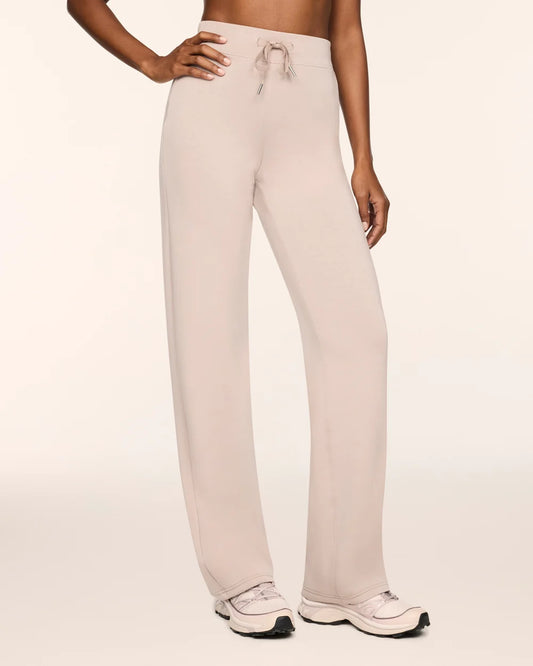 A model wearing light beige wide-leg lounge pants with a drawstring waistband, paired with white sneakers, against a solid off-white studio background.
