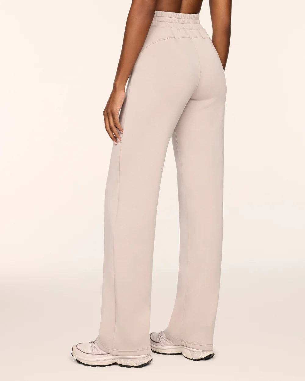 A person shown from the back wearing high-waisted, wide-leg lounge pants in a light warm beige or oatmeal color, paired with white sneakers, against a plain off-white studio background.
