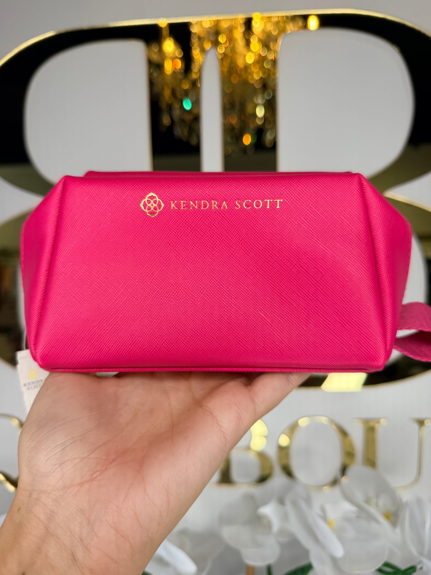 Hand holding a pink pouch with 'Kendra Scott' branding against a blurred decorative background.
