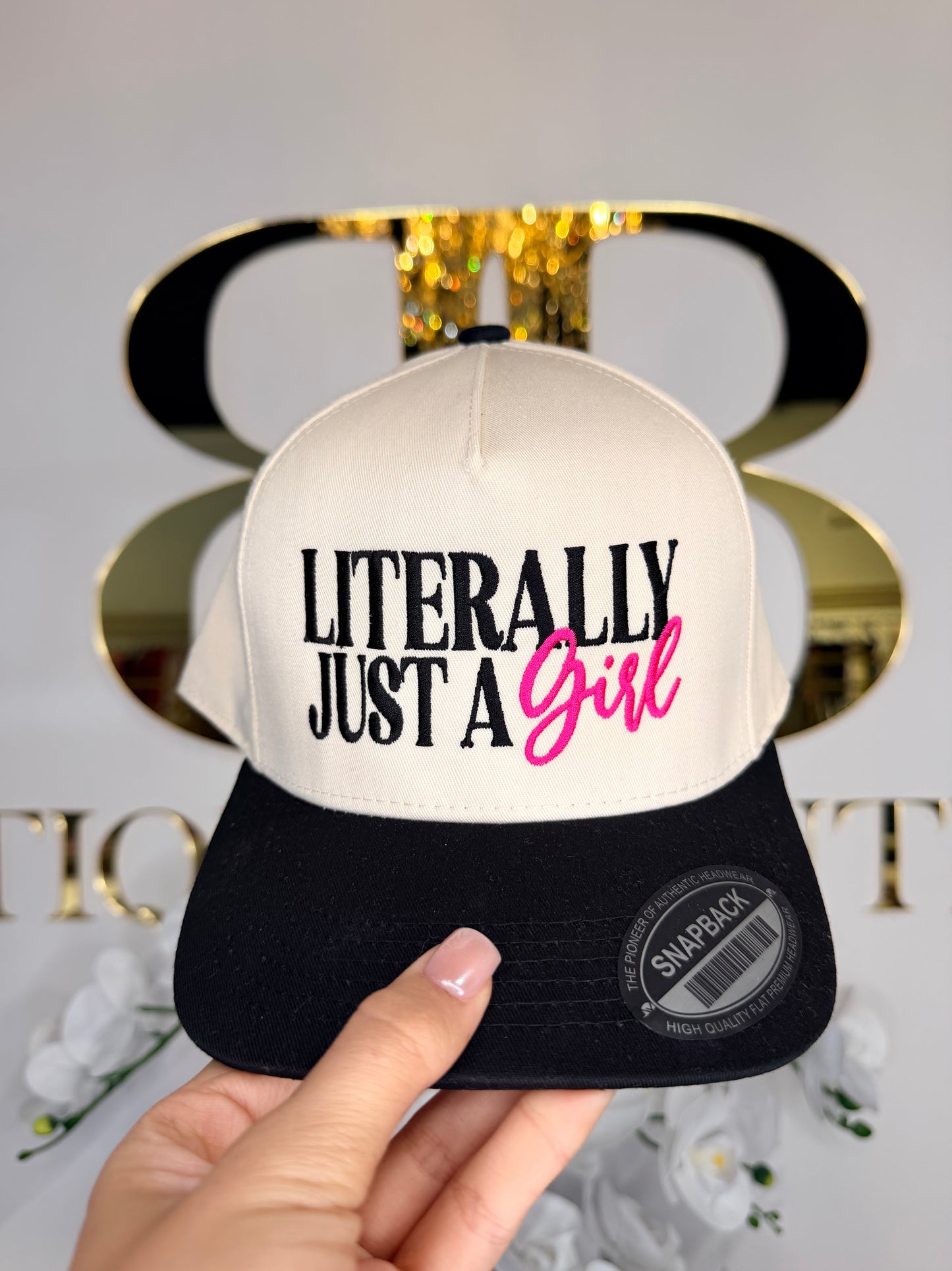Hand holding a cap with 'Literally Just a Girl' text against a blurred background