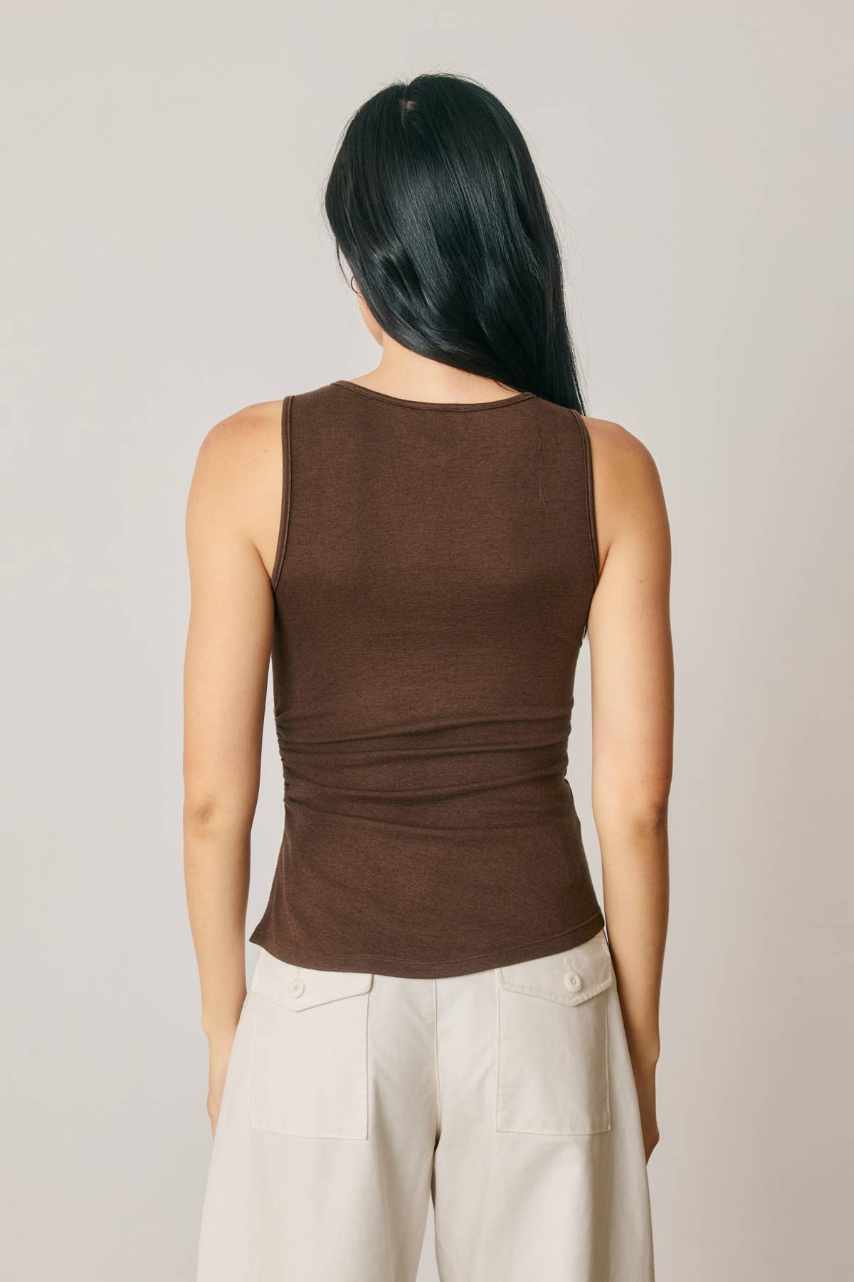 A woman shown from the back wearing a fitted sleeveless brown top and light beige pants against a plain light gray studio background.