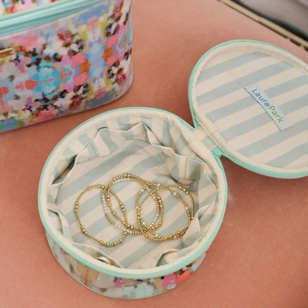 Round jewelry box with striped interior, gold bracelets inside, and a colorful floral case in the background.