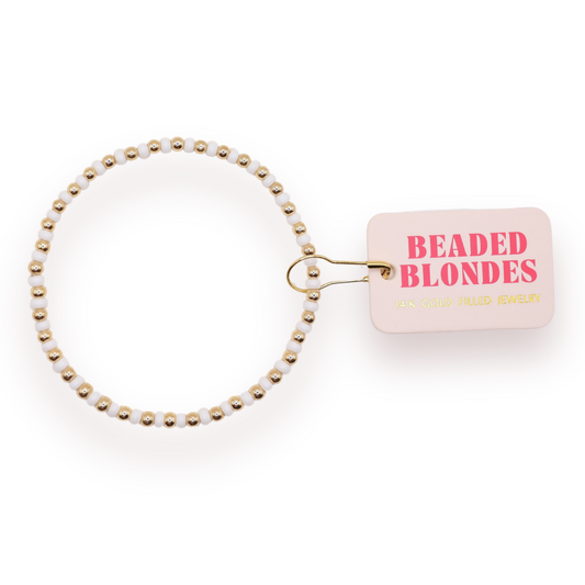 A beaded stretch bracelet with white beads and gold-toned spacer beads, attached to a small pink tag that reads 'BEADED BLONDE JEWELRY' in pink and gold lettering, on a white background.