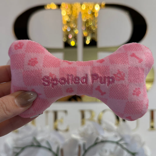 Pink bone-shaped toy with 'Spotted Pup' text held by a hand against a blurred indoor background.