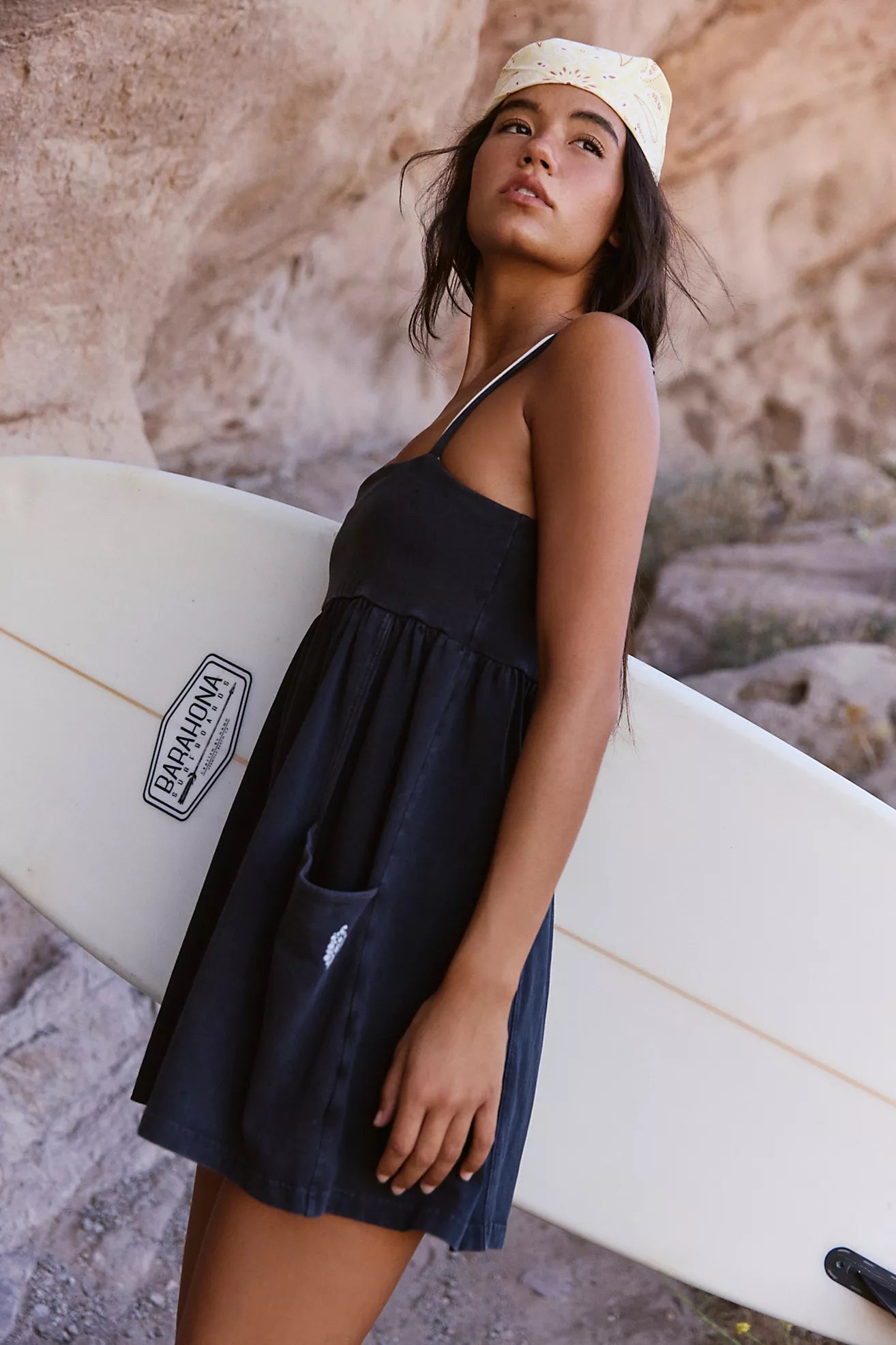 Woman holding a white surfboard with a visible brand logo, standing against a rocky background.