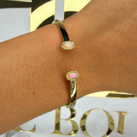Gold bracelet with pink gemstones on a wrist, with a blurred background featuring gold letters and white flowers.