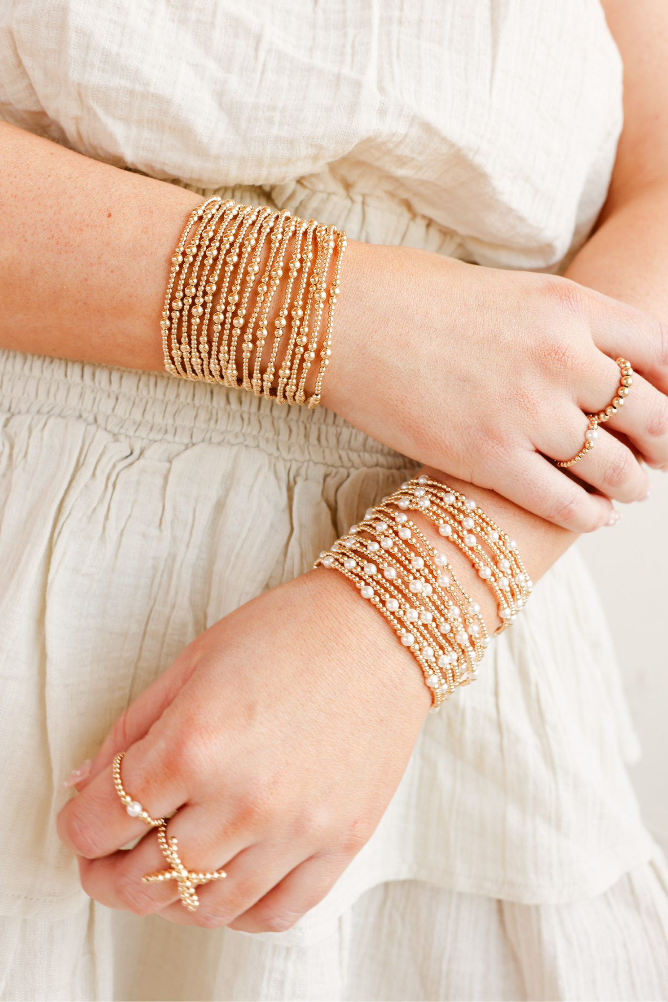 Close-up of hands wearing multiple gold bracelets and rings on a light background