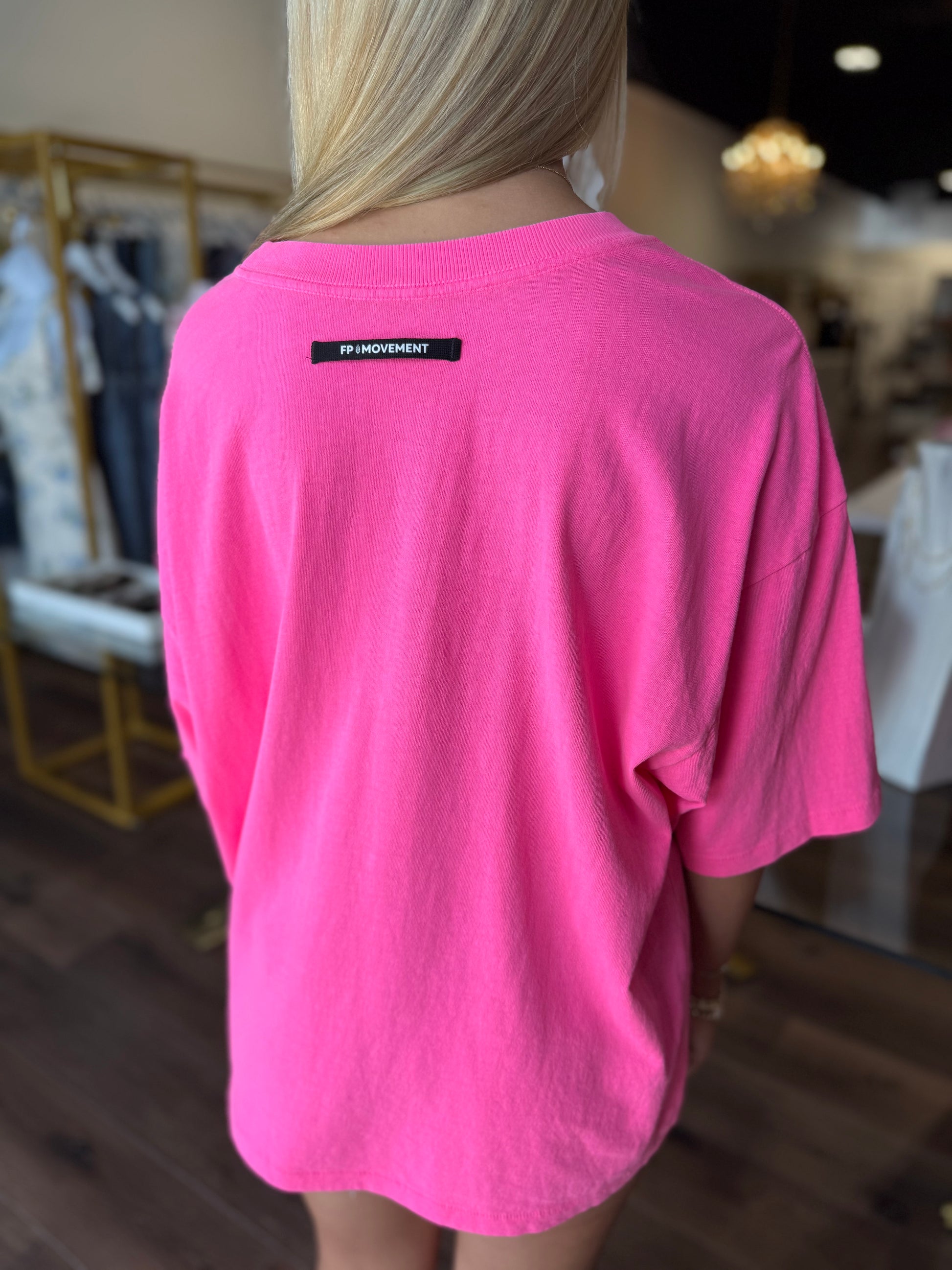 A woman viewed from behind wearing an oversized hot pink t-shirt with a small black rectangular label on the upper back that reads 'F*ck Independence'. The setting appears to be a clothing boutique with wooden floors and racks in the background.