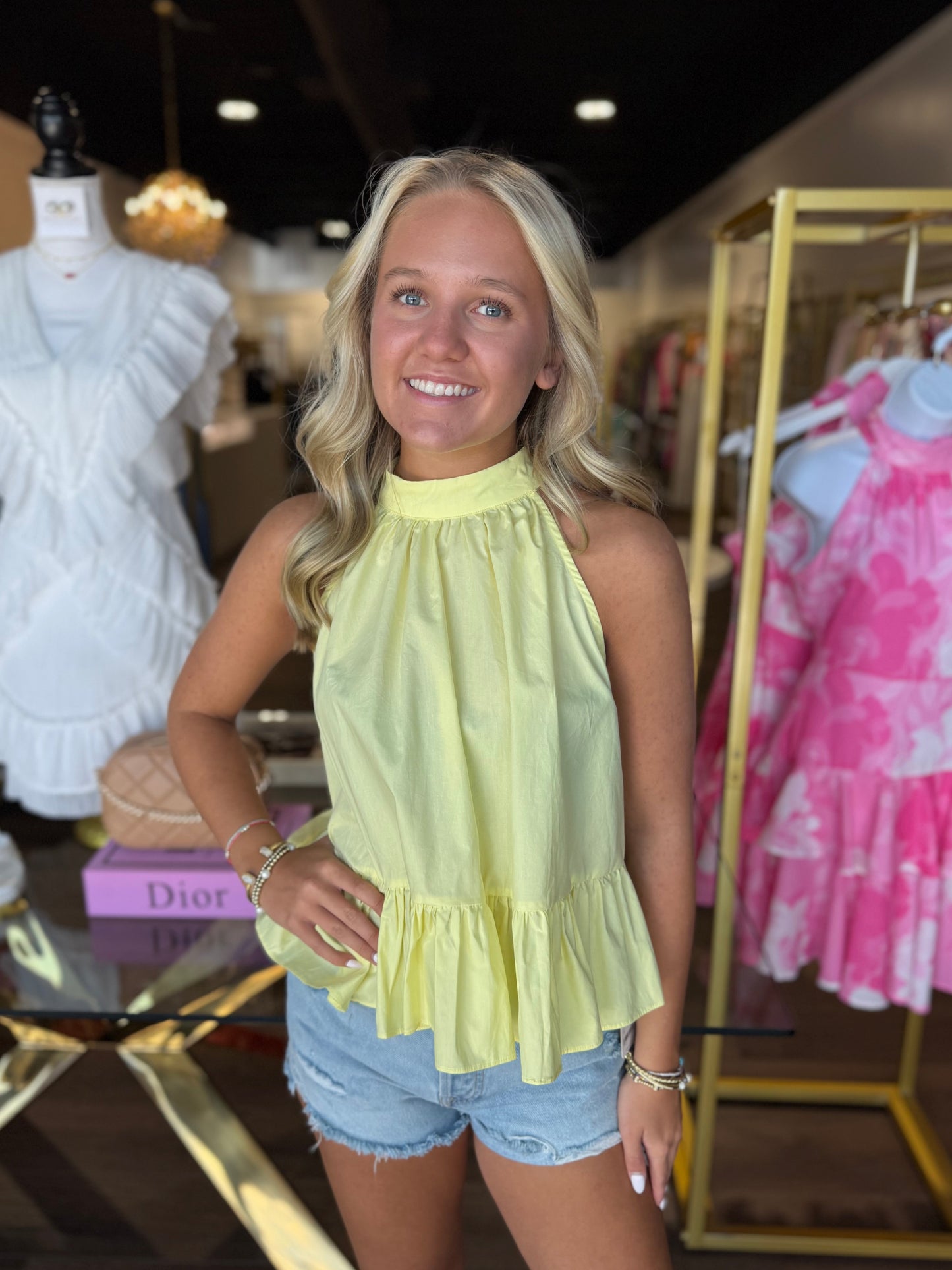 A smiling blonde woman stands in a boutique wearing a pale yellow sleeveless halter top with a ruffled hem and light blue denim shorts. Mannequins and clothing racks are visible in the background.