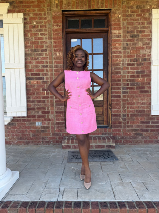 A woman posing on a brick porch wearing a sleeveless hot pink mini dress with gold buttons and beige heels.
