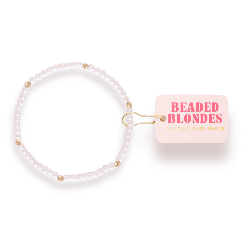 A beaded stretch bracelet made of white pearls with small gold spacer beads, shown next to a light pink tag that reads 'BEADED BLONDES' and 'GOLD FILLED JEWELRY'.