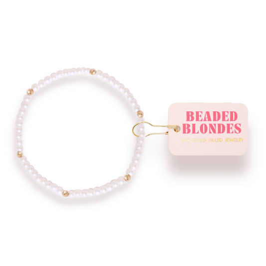 A beaded stretch bracelet made of white pearls with small gold spacer beads, shown next to a light pink tag that reads 'BEADED BLONDES' and 'GOLD FILLED JEWELRY'.