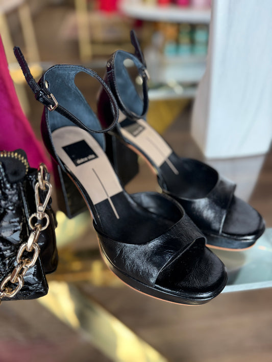 Black Dolce Vita high-heeled sandals on a glass surface with a blurred background