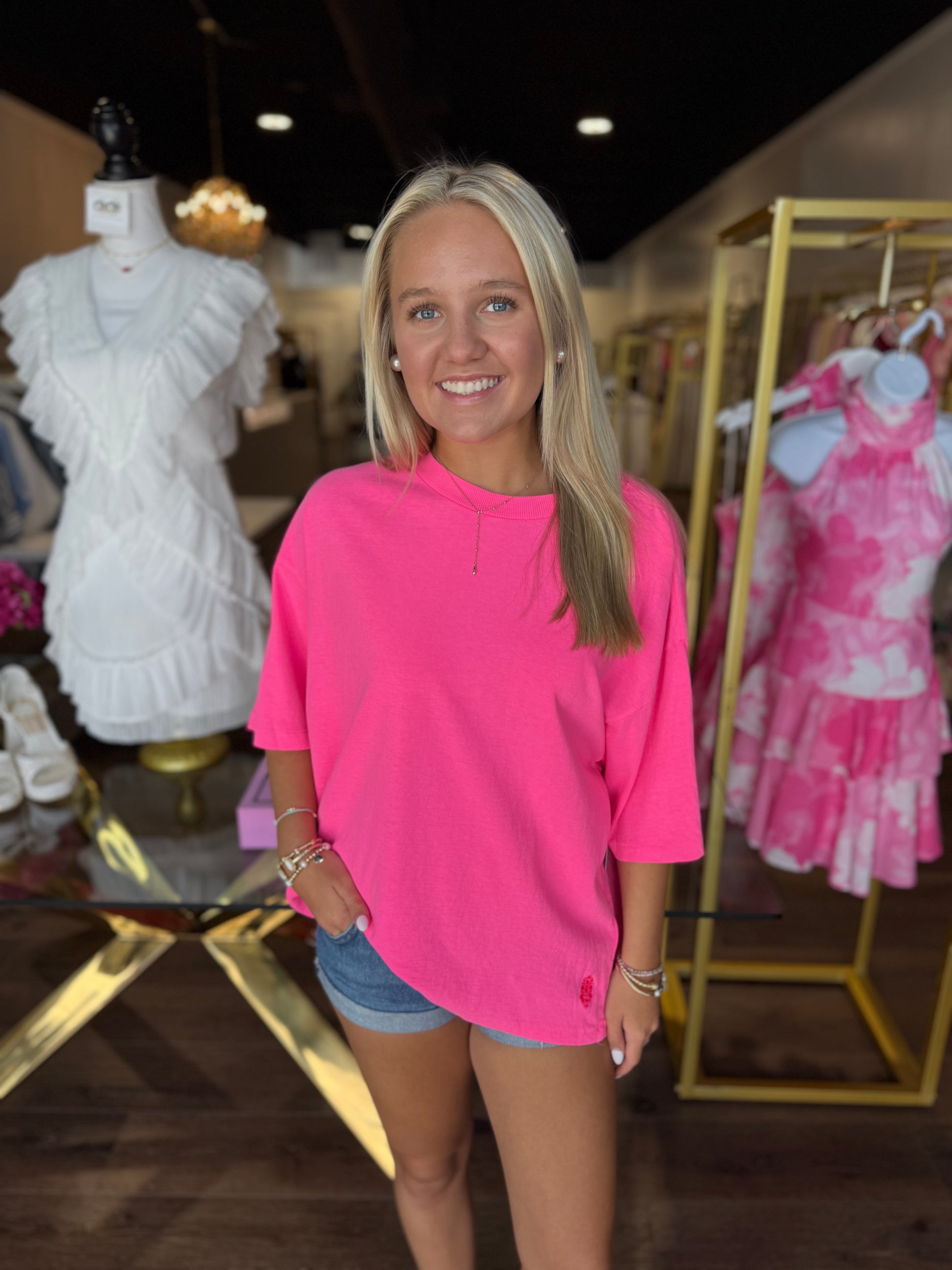 A smiling blonde woman stands in a clothing boutique wearing a bright pink oversized short-sleeve top and denim shorts. Mannequins and racks of clothing are visible in the background.