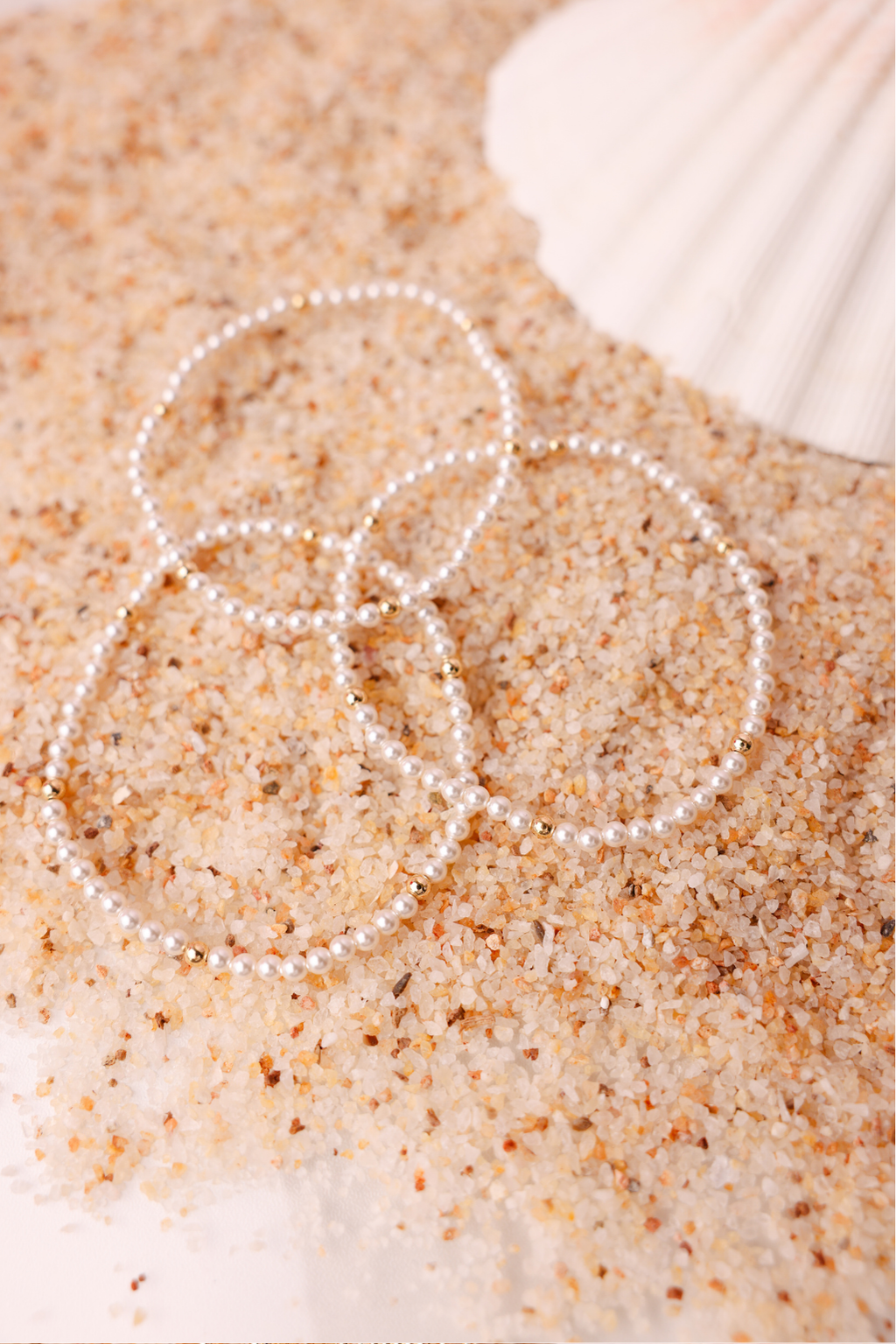 Two pearl bracelets on sand with a shell in the background