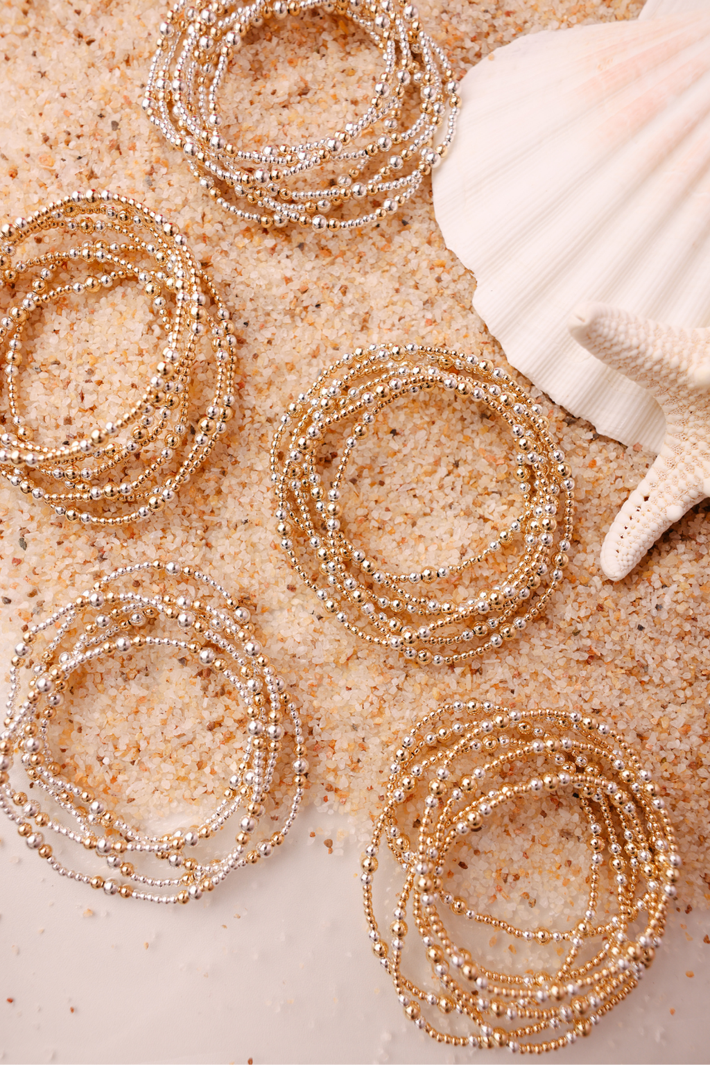 Gold beaded bracelets on sand with a shell