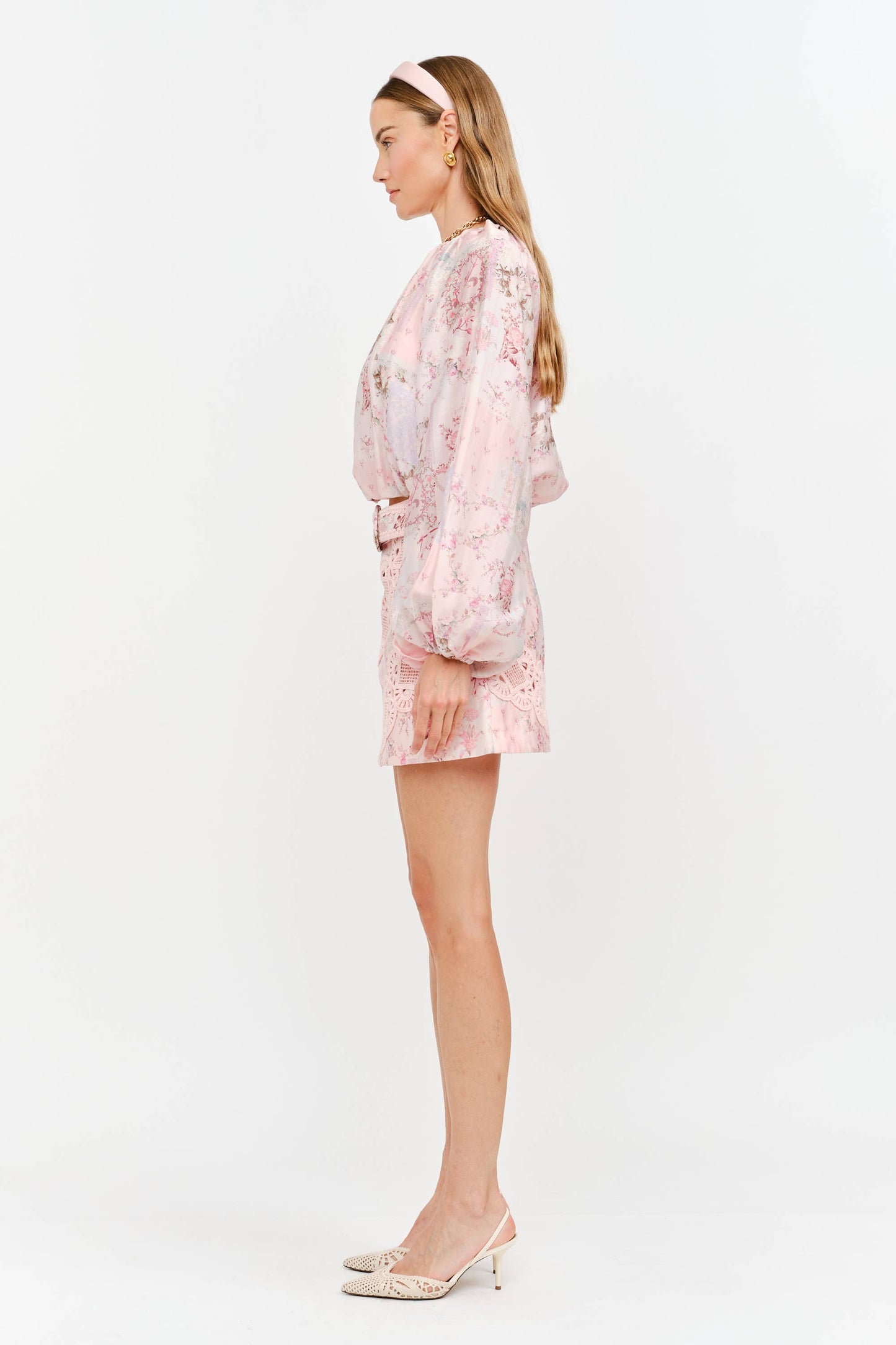 A woman shown in side profile wearing a light pink floral long-sleeve romper with a matching belt, paired with beige pointed-toe heels, against a plain white studio background.