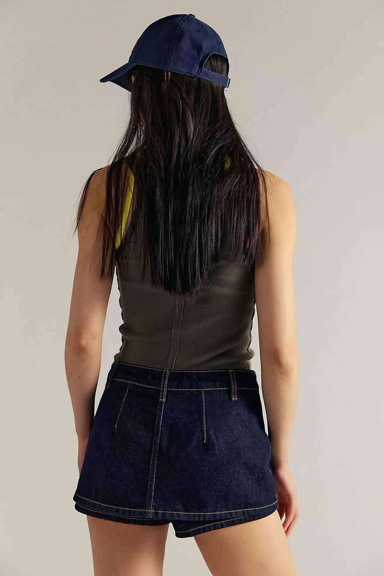 Person wearing a navy cap, green sleeveless top, and dark denim shorts on a plain background