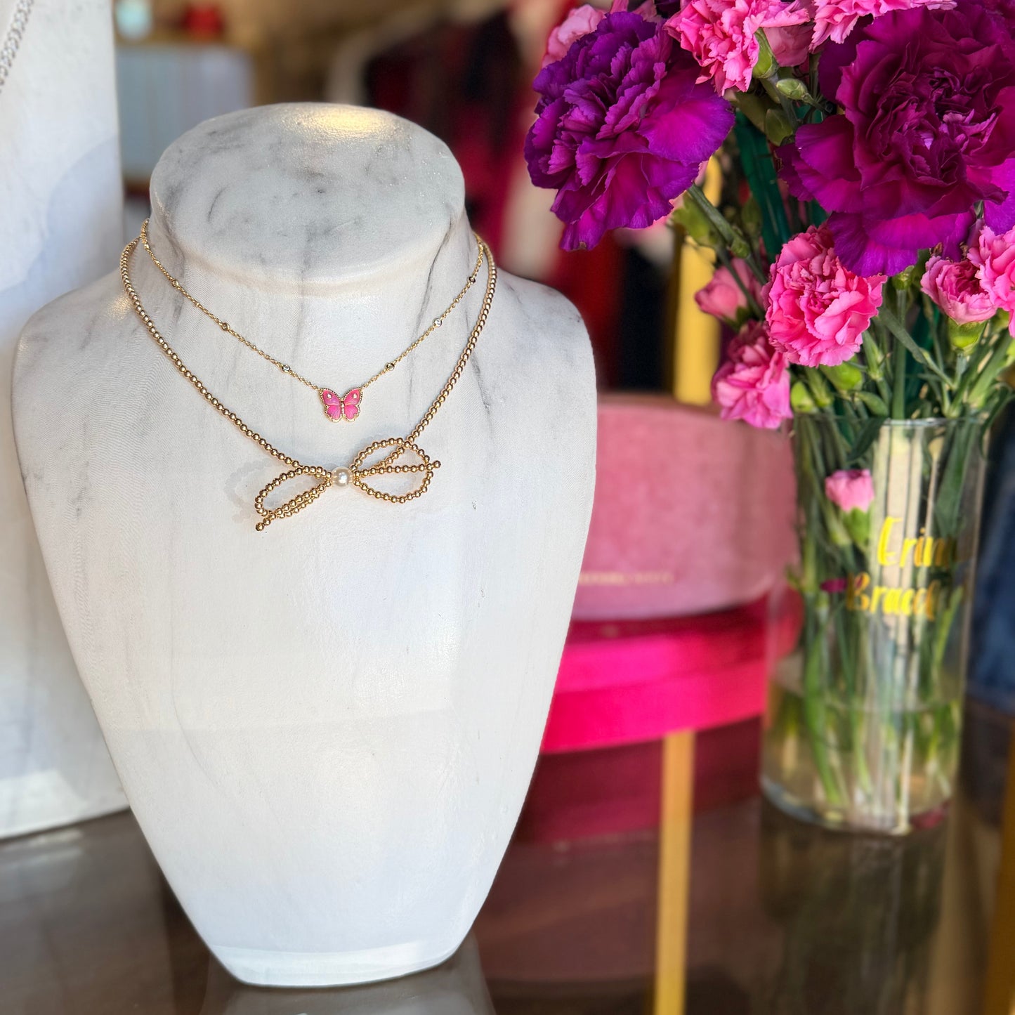 Jewelry display with a Beaded Blondes bow necklace and a Kendra Scott butterfly necklace on a marble bust and a vase of flowers in the background.