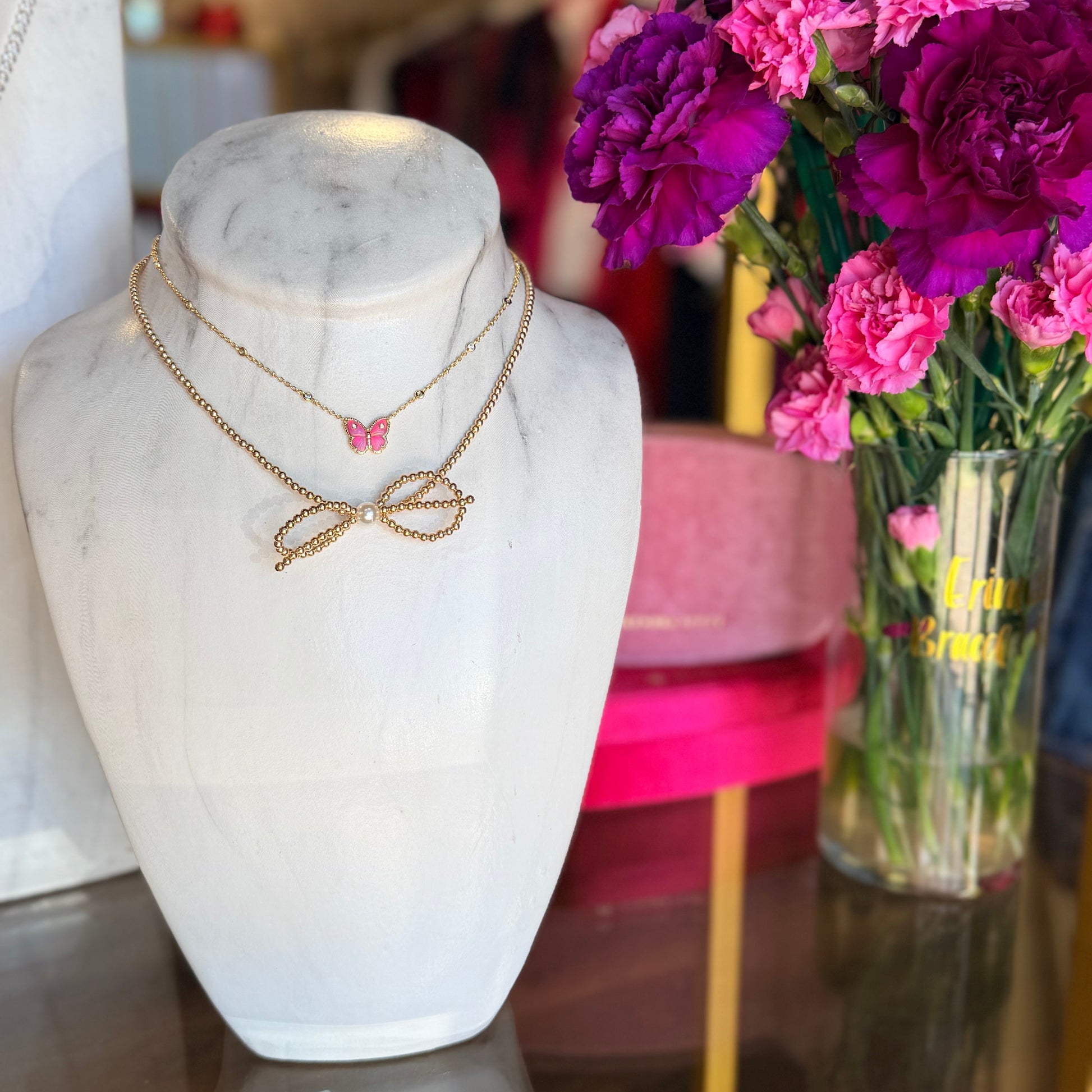 Jewelry display with a Beaded Blondes bow necklace and a Kendra Scott butterfly necklace on a marble bust and a vase of flowers in the background.