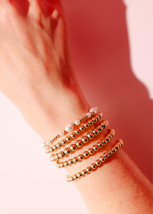 Close-up of a wrist wearing multiple gold beaded bracelets on a pink background