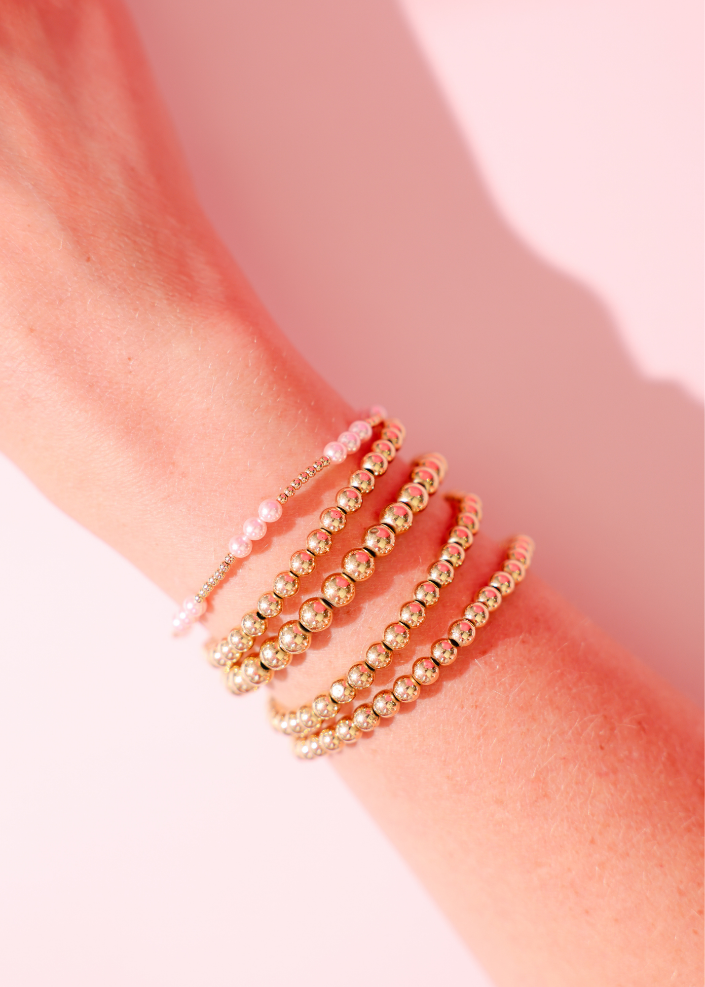 Gold beaded bracelets on a wrist against a pink background
