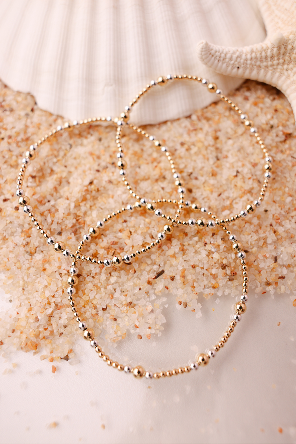 Gold beaded bracelets on a sandy surface with shells in the background