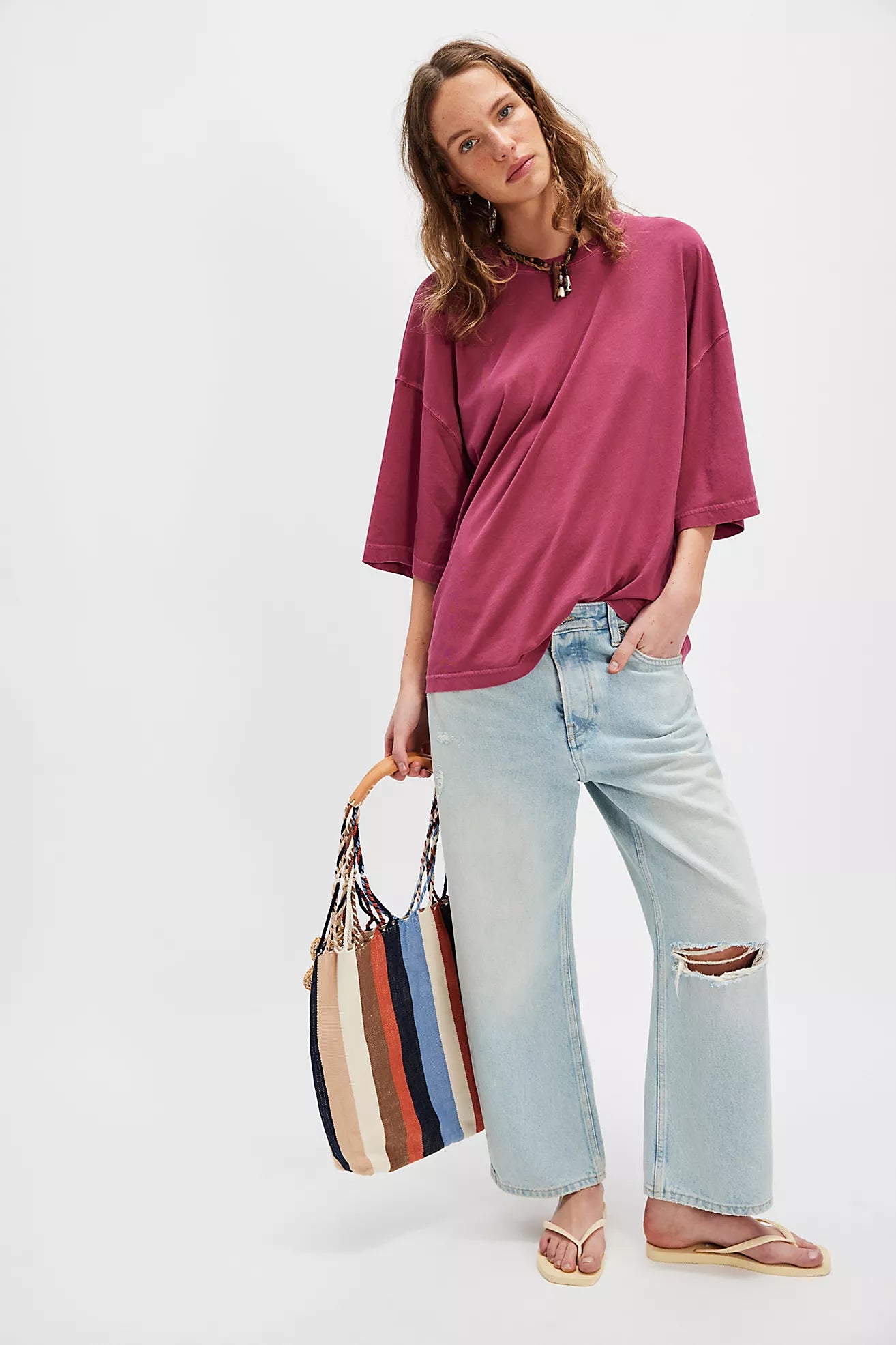Woman wearing a maroon oversized shirt and light blue jeans, holding a striped bag on a white background